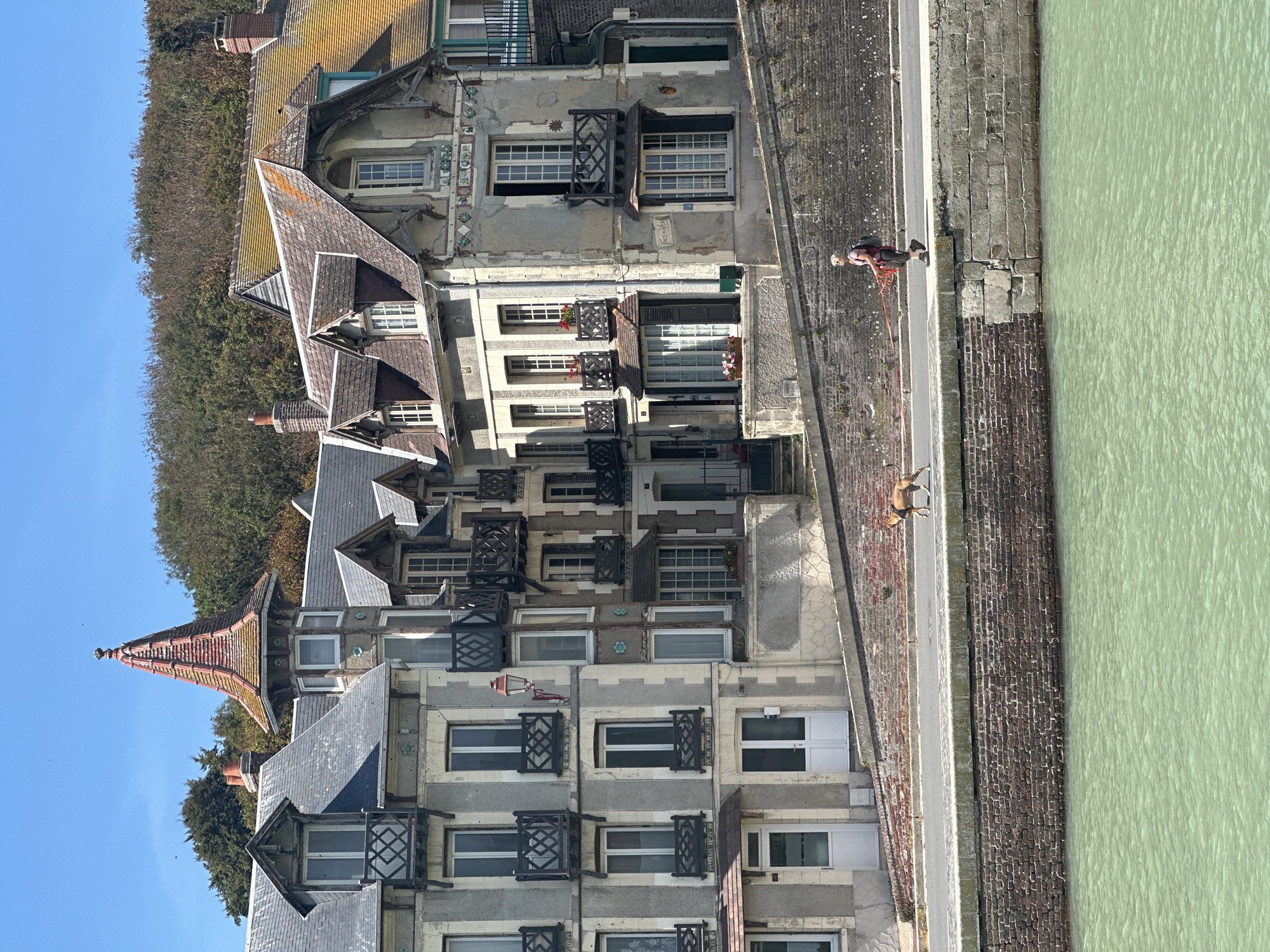 The image depicts a waterfront scene in Saint-Valery-en-Caux, France. In the foreground, a body of greenish water is visible, bordered by a stone embankment and a brick wall. A paved walkway runs along the embankment, where a person is walking a dog. The person, wearing a backpack, is holding a leash attached to the dog. Behind the walkway, a row of multi-story buildings with distinctive architecture lines the waterfront. These buildings feature grey facades, tiled roofs, balconies with intricate railings, and numerous windows. Some of the buildings have decorative elements and dormer windows. The sky above is clear and blue, with some trees visible on a hill in the background. The overall impression is of a sunny day in a historic coastal town.