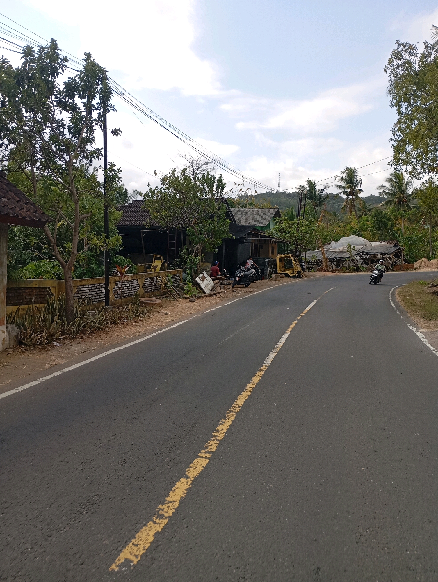 The image depicts a roadside scene in Wonogondo, Indonesia. A paved asphalt road with a broken yellow center line curves through the frame, leading towards a slightly overcast sky with scattered clouds. On the left side of the road, a low yellow wall with a brick pattern separates the road from a residential area. Adjacent to the wall is a tree and a dark-colored house with a tiled roof. Several motorbikes are parked along the roadside in front of the houses. Further down the road, a motorcycle is seen in motion, riding away from the camera. The environment is characterized by lush green vegetation, palm trees, and some scattered debris, suggesting a rural or semi-urban setting. The overall impression is a slice of everyday life on a rural road in Indonesia.