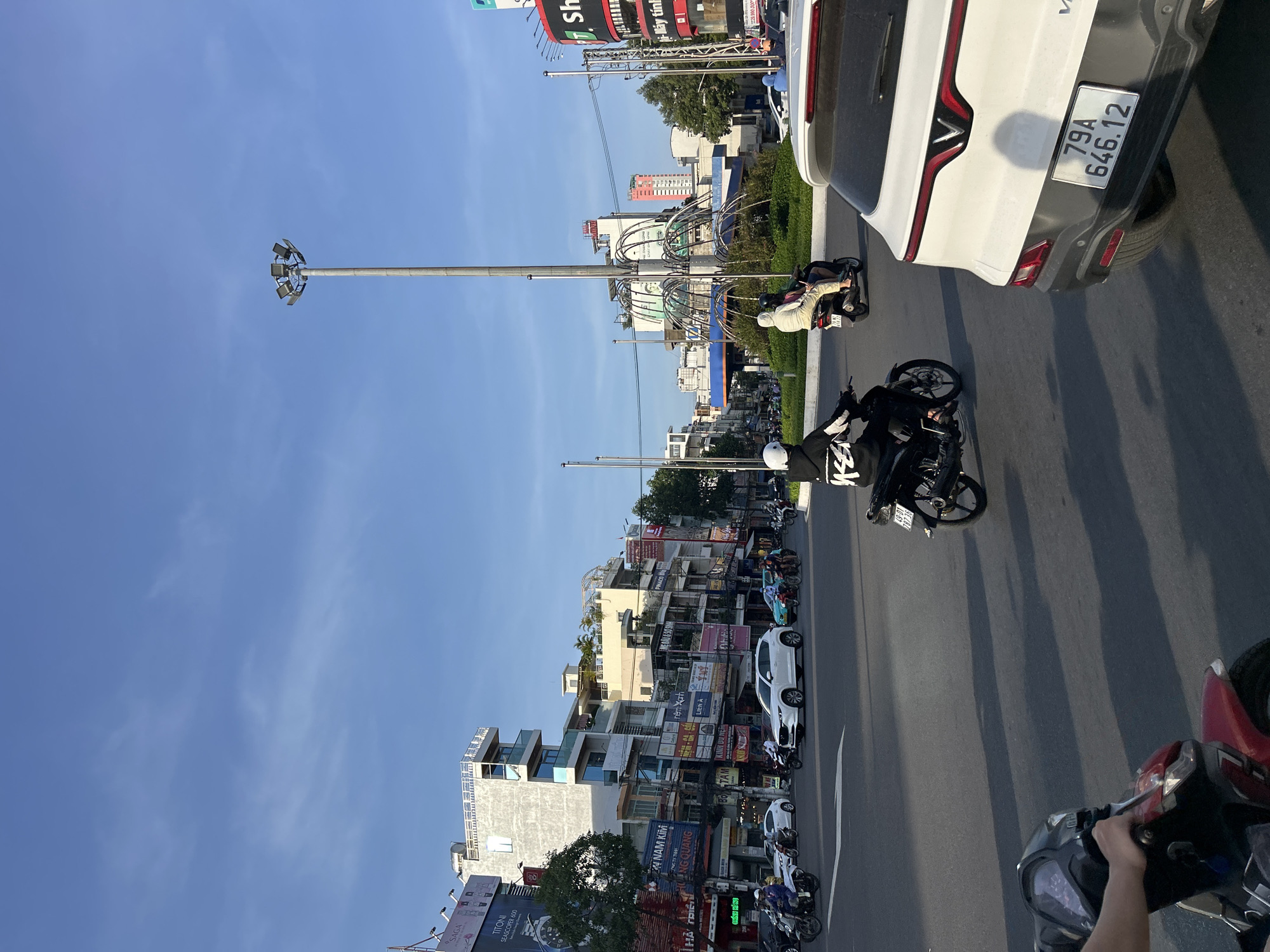 The image captures a street scene in Nha Trang Ward, Vietnam, characterized by a mix of traffic, commercial buildings, and urban infrastructure. A prominent feature is a large, multi-head street lamp positioned centrally, illuminating the intersection. Several vehicles, primarily motorcycles with riders, are navigating the roadway. A white SUV, its rear prominently displayed, is parked or moving in the foreground, displaying a Vietnamese license plate. In the background, a cluster of multi-story buildings with various signage and architectural styles line the street, suggesting a commercial district. The sky is clear and blue, indicating daytime. A circular fountain structure is visible behind the street lamp, surrounded by greenery. The overall impression is of a busy urban environment during daylight hours.