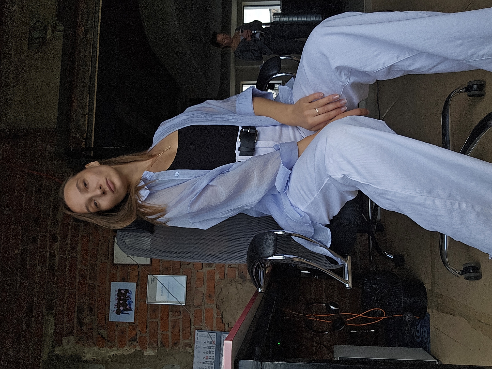 A woman with long brown hair sits in an office chair in a room with exposed brick walls. She is wearing a light blue unbuttoned shirt over a black tank top and white pants. Her hands rest on her lap, and she wears a ring on her right ring finger. In the background, another woman stands by a window, looking at her phone. A desk with headphones, a trash can, and framed pictures on the wall are visible. The room appears to be a modern office space, possibly in an industrial building. There is no information available to confirm the location as Chaikovsky, Russia.