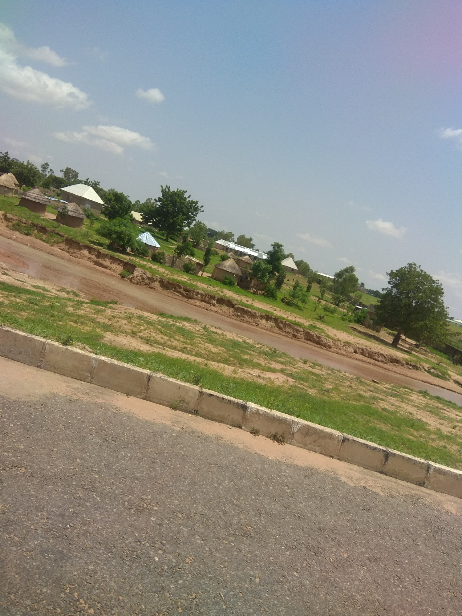 The image depicts a rural village scene, likely in Jalingo, Nigeria. In the foreground, a paved road with a concrete curb runs diagonally across the frame. The road appears to be somewhat worn. Beyond the road, a slope covered with dry grass and some green patches leads up to a cluster of traditional thatched-roof huts and a few buildings with more modern, angular roofs. Some of these structures appear to be homes, while others might be community buildings or agricultural facilities. The landscape is dotted with various trees, some with full green foliage and others appearing more sparse. In the background, the sky is a pale blue with scattered white clouds, suggesting a clear day. The overall impression is one of a developing community in a dry, perhaps semi-arid, environment. There are no people visible in the image.