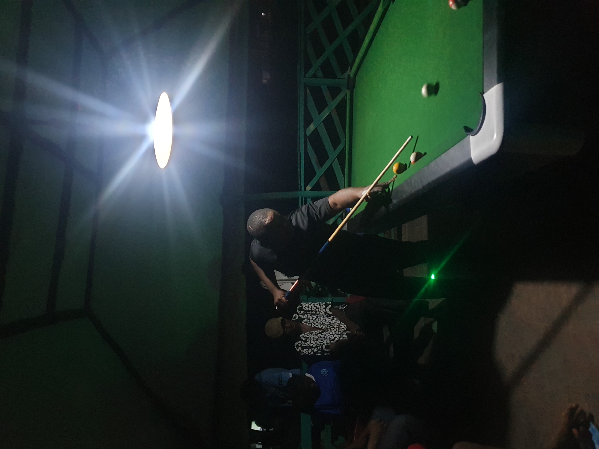 The image depicts an outdoor gathering at night, likely in Lokoja, Nigeria. In the foreground, a person is actively playing pool on a green felt table, using a cue stick to strike a ball. Several other pool balls are visible on the table. To the left of the pool table, a group of individuals are seated, some appearing to be observing the game. The environment is dimly lit by a bright overhead light, casting strong beams. A green railing with a lattice design encloses the area. The overall setting suggests a recreational space, possibly a bar or a private outdoor entertainment area.