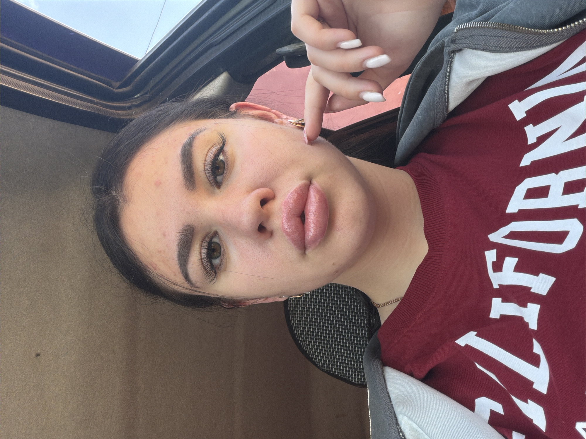 The image is a close-up, selfie-style shot of a young woman in a car. She is wearing a dark red sweatshirt with white lettering that appears to spell "CALIFORNIA". She has dark hair, prominent eyelashes, and full lips. Her right hand is visible, with white painted fingernails, and her index finger is touching her cheek. The interior of the car is visible, including the headrest of a seat and part of the car's door frame. The background is slightly out of focus, showing the car's interior and a hint of the sky outside. There is no indication of the city of Фоки, Russia within the image itself.