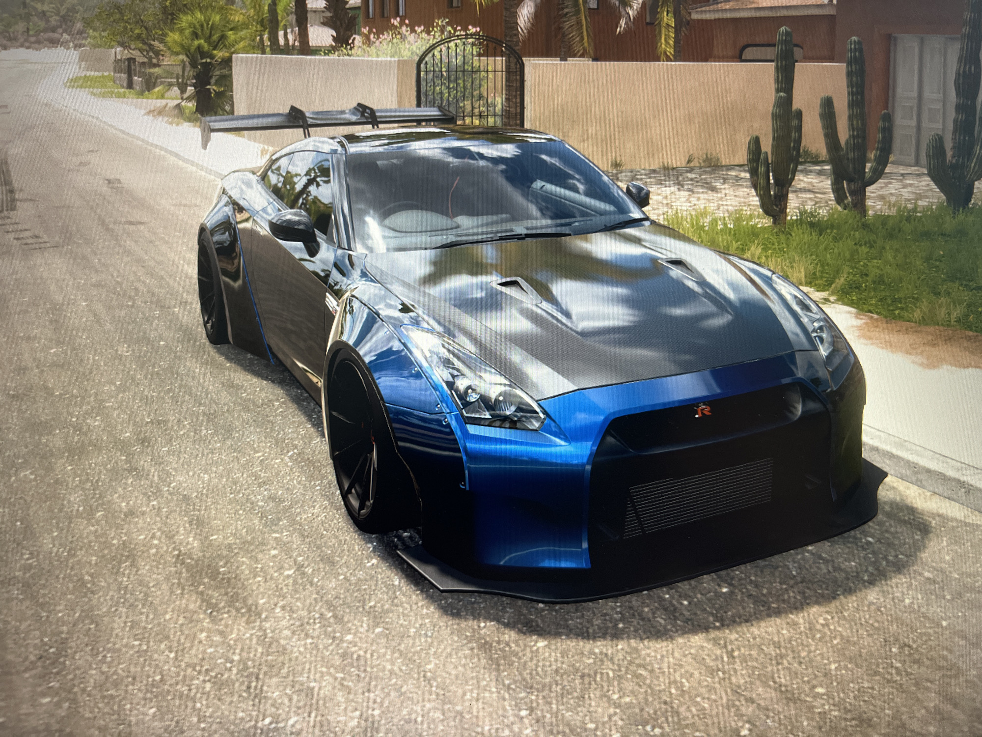 A blue and black Nissan GT-R is parked on a paved street in front of a building with a fence and cacti. The car has a large rear spoiler and a carbon fiber hood. The street is a light gray asphalt with visible texture. The background includes a tan wall, green grass, and palm trees. The scene appears to be a still image from a video game. There are no people or animals visible.
