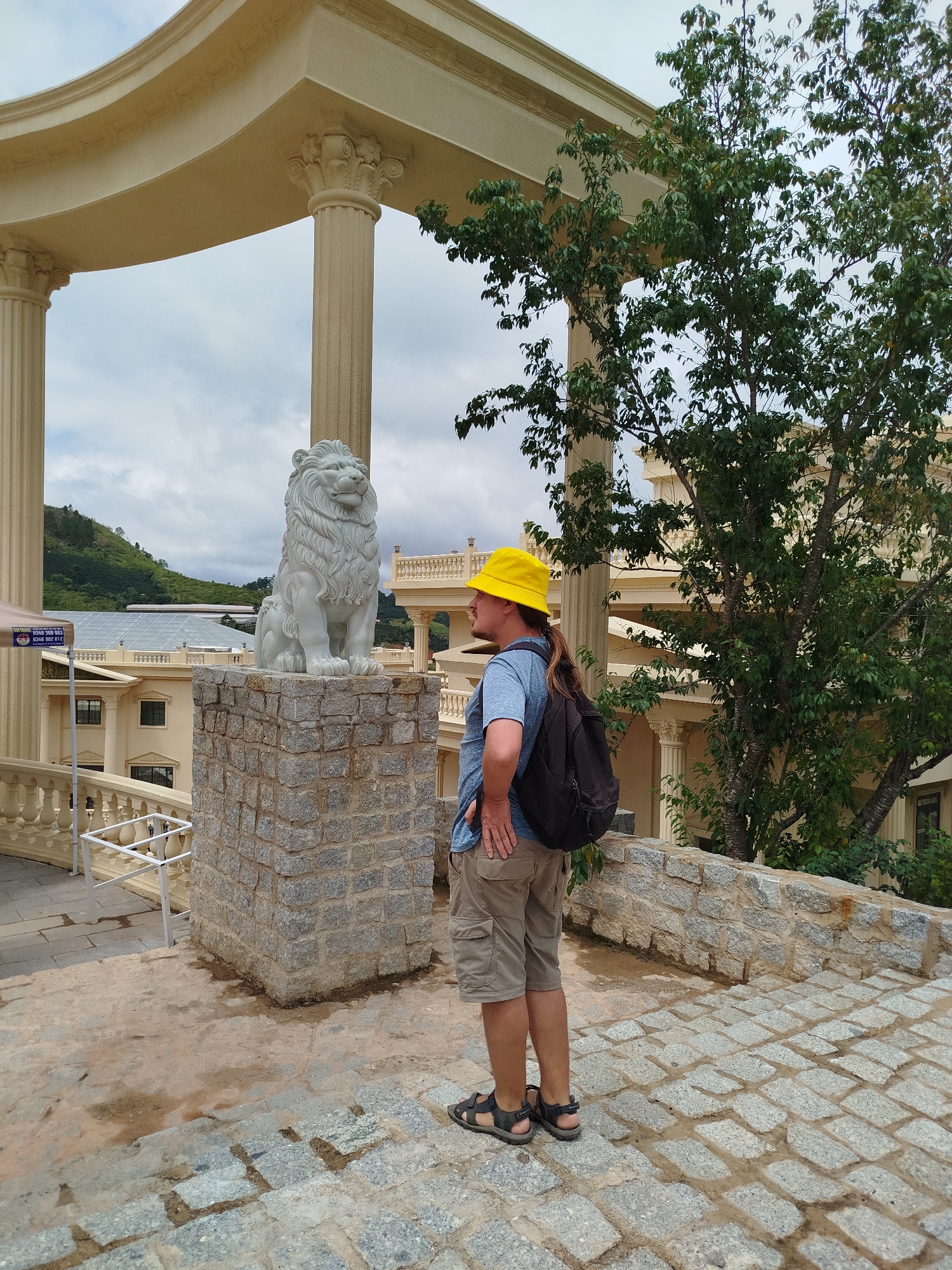A person wearing a yellow bucket hat, blue t-shirt, and khaki shorts stands on a stone-paved path, looking at a white lion statue. The statue is mounted on a stone pedestal. The background features a building with classical architecture, including large columns and an ornate roof. Trees with green foliage are visible, and the sky is overcast. The location is identified as Lạc Dương, Vietnam.