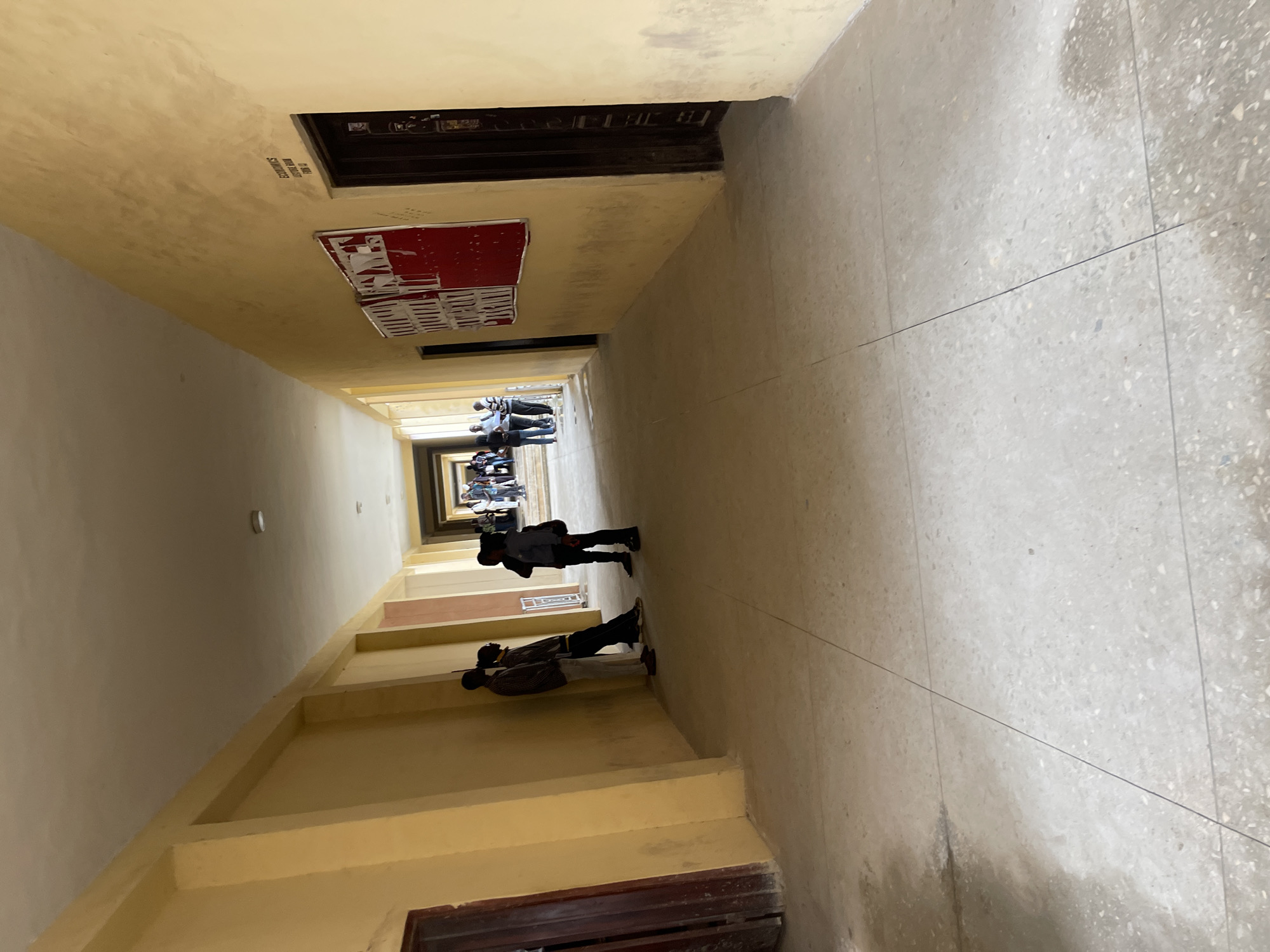 The image depicts a narrow, vertical indoor hallway with a high ceiling, likely part of an apartment building or similar structure in Jos, Nigeria. The hallway is painted a pale yellow, with stark, unadorned walls. The floor is tiled with a speckled, light-colored material, marked by dark grout lines.
Several people are visible, seen from the waist down, walking in both directions along the hallway, their figures diminishing in the distance. They are dressed in casual clothing. In the foreground, two individuals stand facing the camera. One person is wearing a dark jacket and black pants, while the other is in a patterned shirt and dark pants. Further down the hallway, a group of people are gathered, suggesting a communal or social interaction.
The lighting suggests it is daytime, with a soft, ambient light illuminating the space, possibly from a skylight or windows at the far end of the hallway, which are not visible. There are no obvious signs of the weather.
Notable details include a red poster or notice tacked to the wall on the left, partially torn and peeling. Above the poster, a dark wooden doorway is visible, suggesting access to rooms or apartments. Towards the far end of the hallway, the architecture creates a sense of depth and perspective, with multiple doorways and receding walls. The overall impression is one of everyday life in a residential building. No discernible text is legible from the image.
