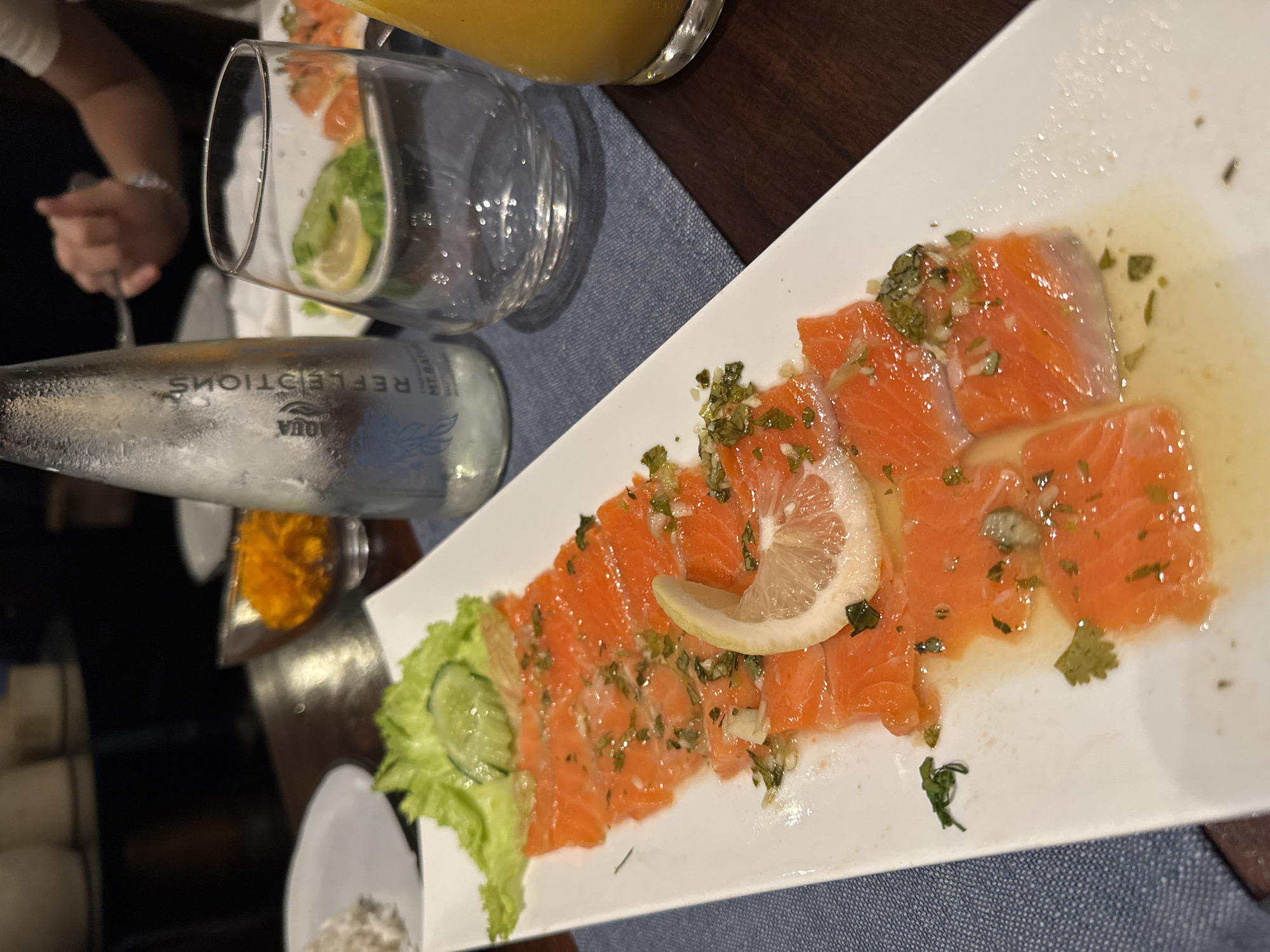 A white rectangular plate of thinly sliced raw salmon, seasoned with herbs and lemon, is presented on a dark wooden table. The salmon is arranged in overlapping rows, garnished with a lemon wedge, lettuce, and cucumber slices. To the right of the plate, a clear glass contains ice and a slice of lemon. Behind the glass, a partially obscured bottle of "Aqua Reflections" mineral water stands, its label visible. A glass of orange juice is also visible to the far right. In the background, a person's hand is visible holding a fork over a plate, suggesting a dining setting. The overall scene appears to be a restaurant in North Kuta, Indonesia, as indicated by the presence of "Aqua Reflections" mineral water, a popular brand in the region. The lighting is dim, characteristic of an indoor evening meal.