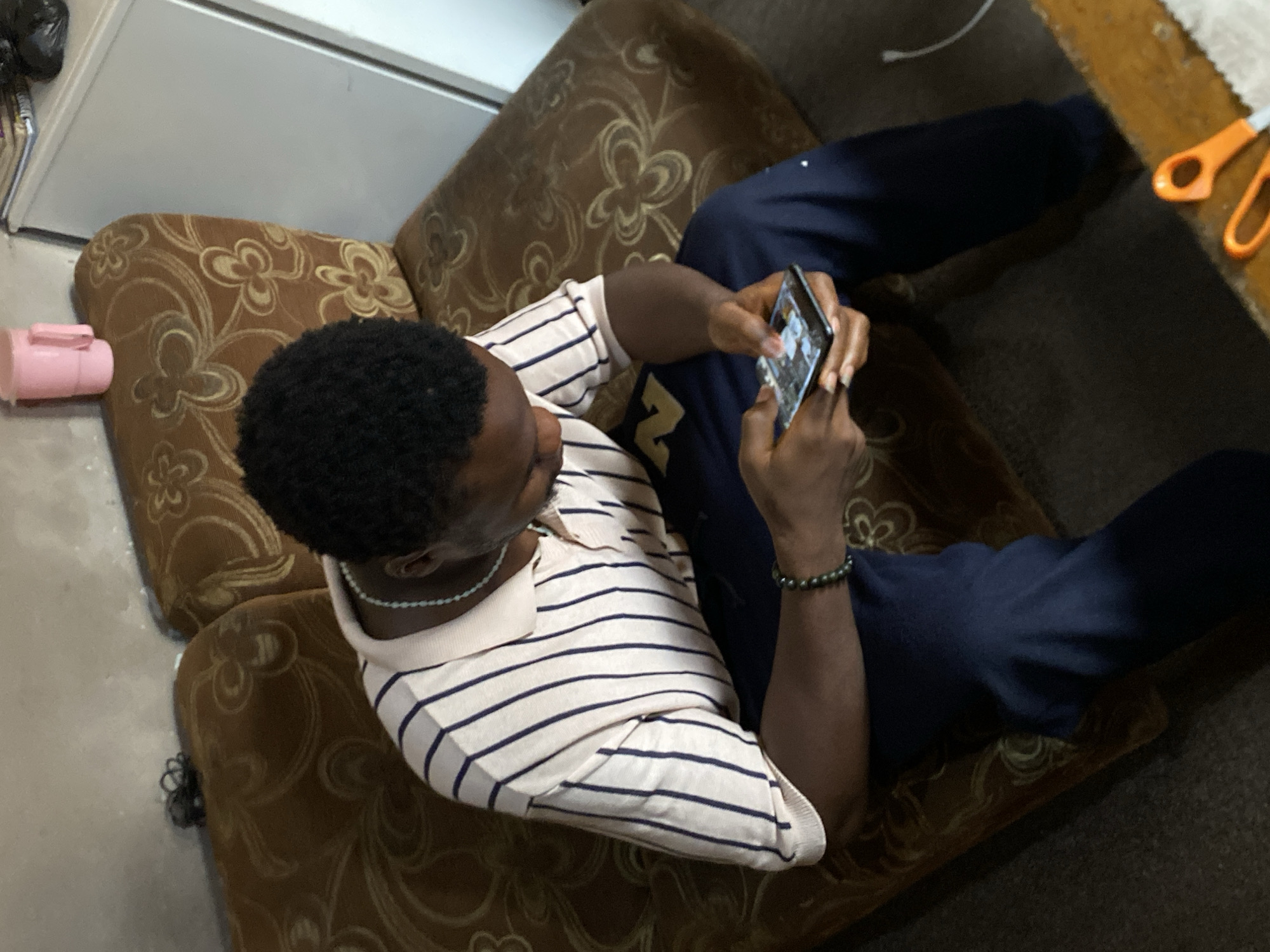 A man is seated on a brown patterned sofa, wearing a striped shirt and dark blue pants. He is holding a smartphone and looking at the screen. The location is Bukuru, Nigeria. In the background, there is a white appliance, a pink object, and an orange pair of scissors on a wooden surface. The floor is dark-colored.