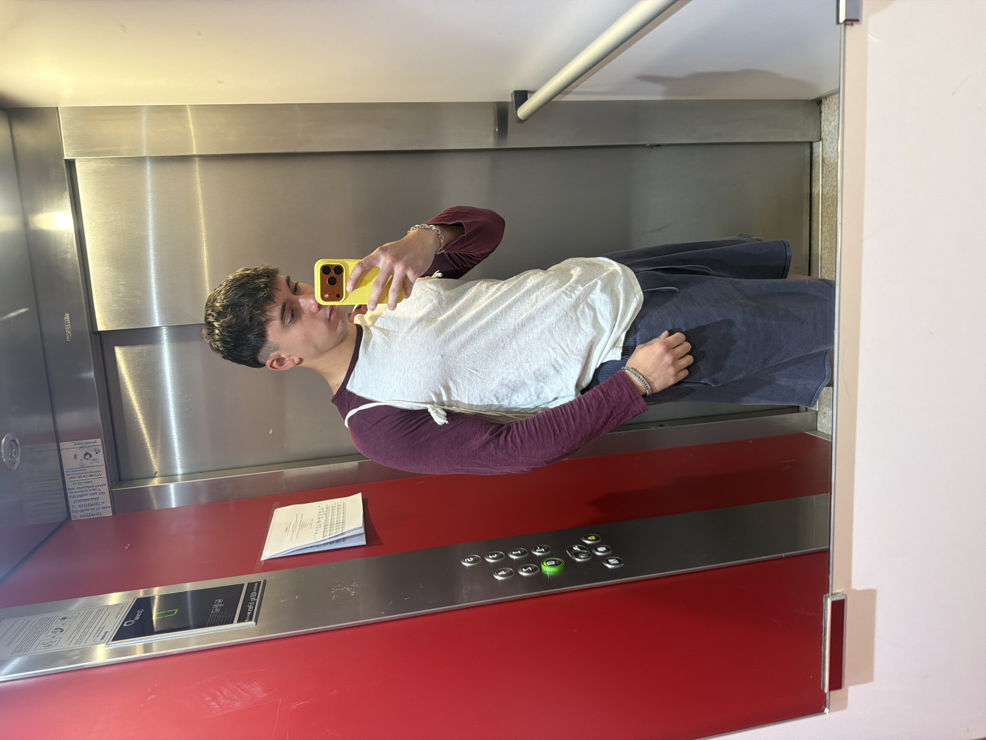 The image depicts a young man taking a selfie in an elevator. The elevator has stainless steel walls and a red lower panel with a control panel. Papers rest on the panel below the control buttons. A handrail is visible at the top of the elevator. The man is wearing a burgundy long-sleeved shirt with a white t-shirt over it and dark-colored shorts or pants. He is holding a yellow smartphone with multiple camera lenses. There is no explicit evidence in the image to confirm its location in Sofia, Bulgaria.