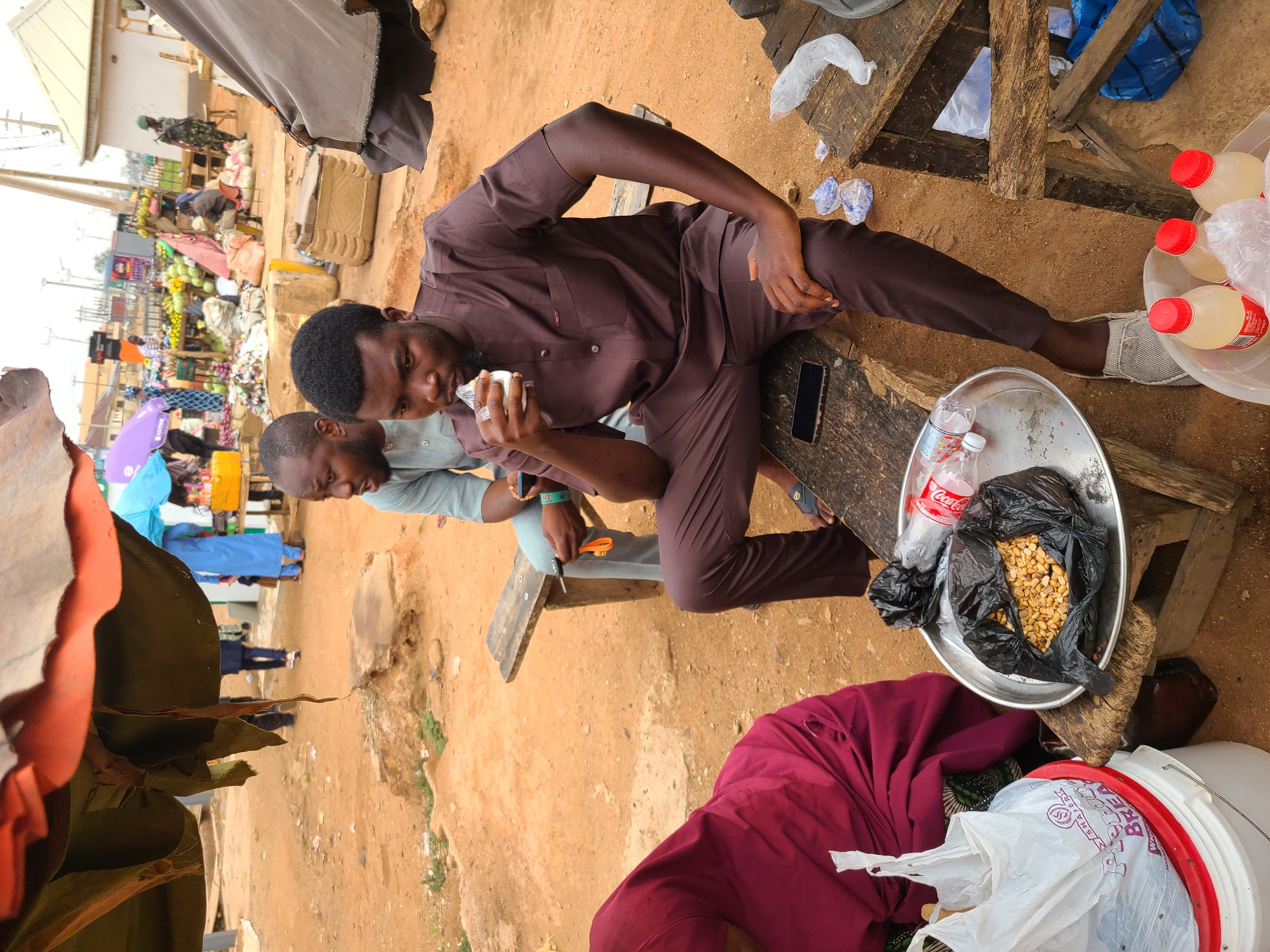 The image shows two men sitting on a makeshift wooden bench in what appears to be an outdoor market setting in Jos, Nigeria. The man on the right is wearing a brown long-sleeved shirt and pants, and he is holding a small, clear plastic container to his mouth. His cell phone sits on the bench nearby. To his right is a metal tray holding peanuts in a black plastic bag and a plastic Coca-Cola bottle. Next to the tray sits a plastic bucket. The man to the left is wearing a light blue shirt and is holding a small orange tool. A crowded market scene is visible in the background. The ground is dirt.