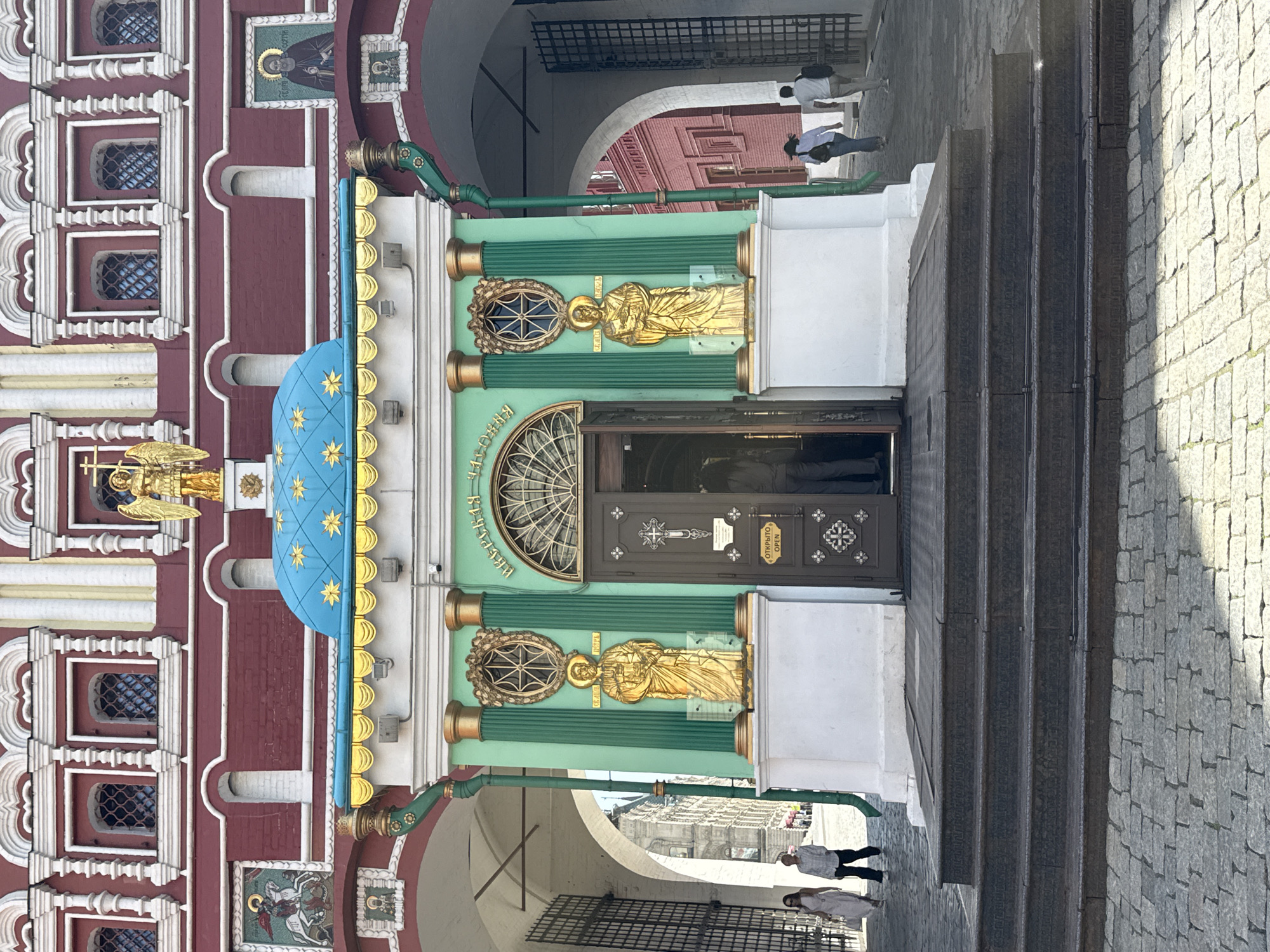 The image shows the entrance of the Kazan Cathedral in Moscow, Russia. The entrance features a green facade adorned with golden statues and a semi-circular window above the door. A blue dome with gold stars is adjacent to the entrance. The building is located at the Red Square in Moscow. There are steps leading up to the entrance. Pedestrians can be seen walking near the building. The surrounding area is paved with stone blocks.