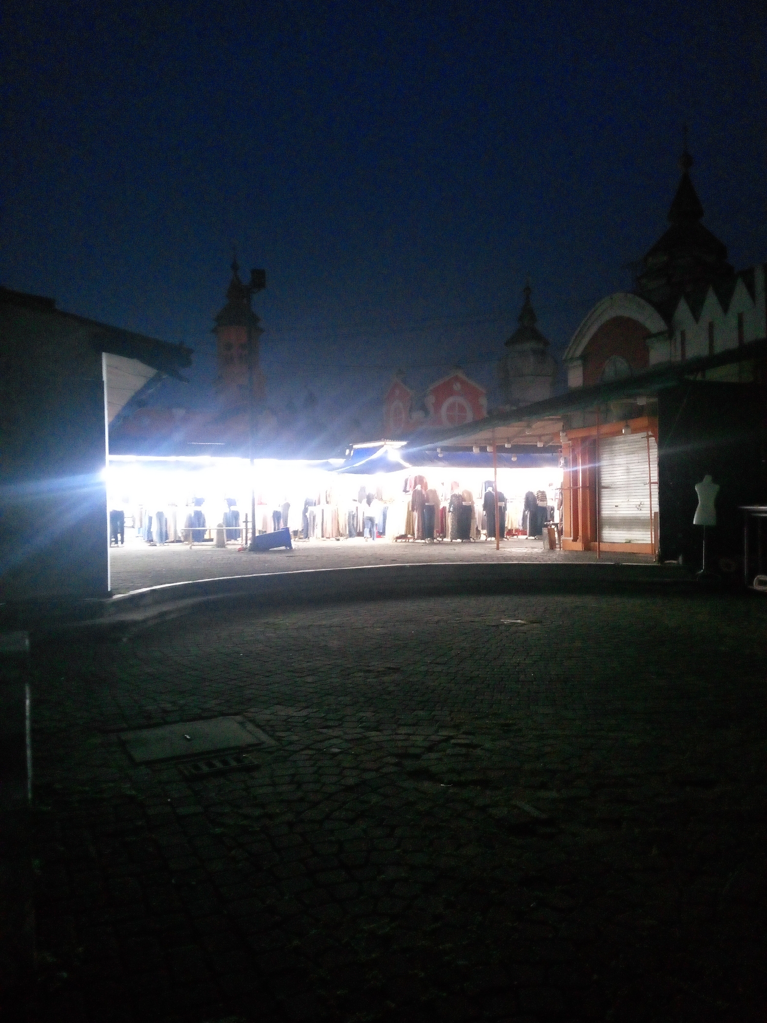 The image depicts a nighttime scene at an outdoor market, possibly a street market or bazaar, illuminated by bright artificial lights. Several stalls are visible, displaying various goods, predominantly clothing, which are hung on racks. A mannequin is present outside one of the stalls. The ground is paved with cobblestones. In the background, there are architectural structures with distinct spires and rooflines, suggesting a location with historical or religious significance. The sky is dark blue. The image quality is poor, with a lot of noise and low resolution, making it difficult to discern finer details. The scene appears to be deserted, with no people actively engaged in shopping or selling. The presence of bright lights and neatly displayed goods indicates that the market is operational or has recently been so. The overall impression is of a quiet, possibly post-business or pre-opening, market setting at night.