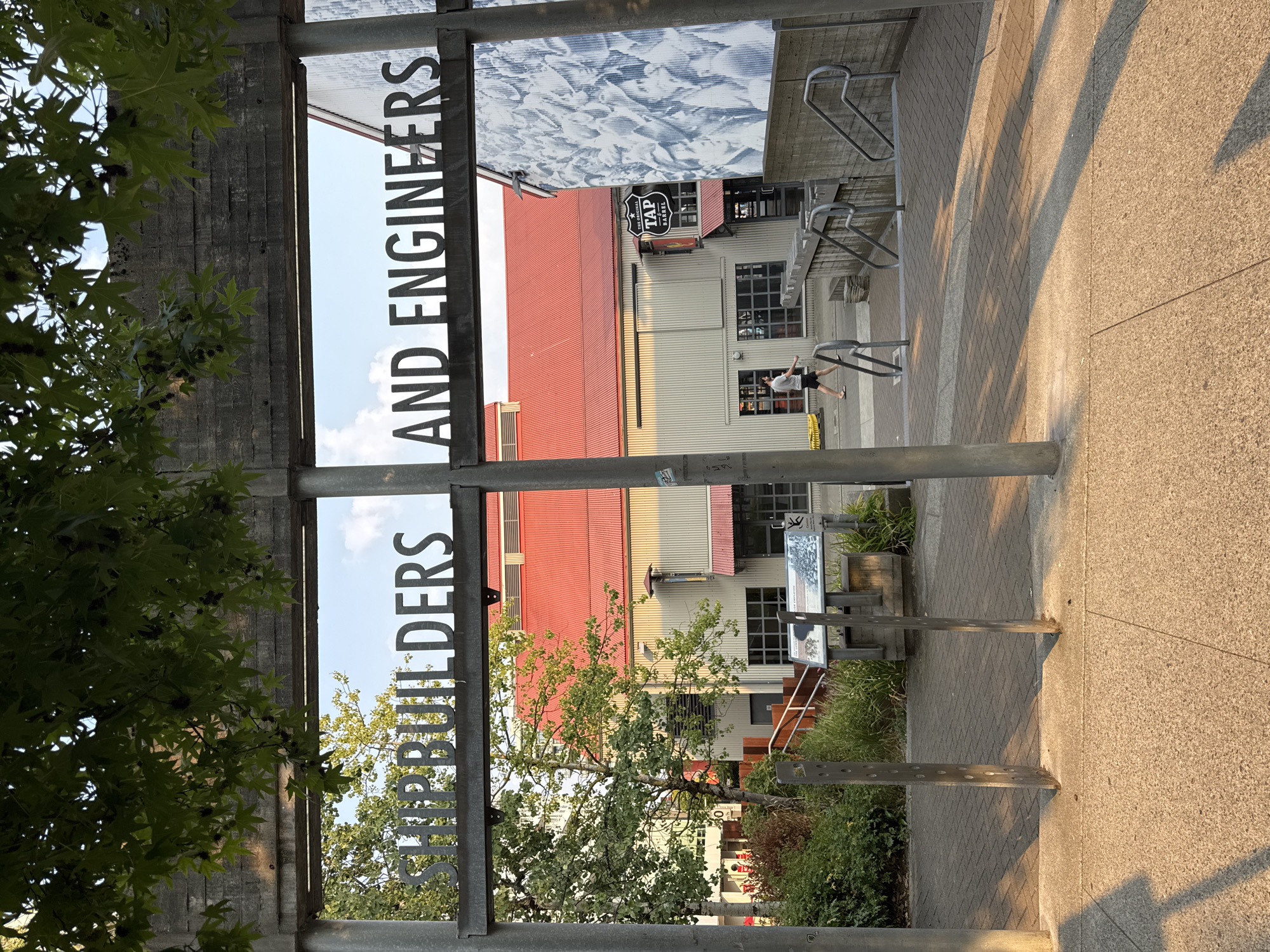 An exterior, eye-level, medium shot captures a gateway entrance to a building complex in North Vancouver, Canada, at what appears to be late afternoon. Metal letters forming the words "SHIPBUILDERS AND ENGINEERS" are mounted horizontally on a concrete beam, which is supported by two vertical metal posts. Beneath this archway, a paved walkway leads toward a modern building with a corrugated metal exterior and a bright red roof. To the right of the building, a large mural depicting faces is visible on another structure. A single individual, wearing shorts and a t-shirt, walks away from the camera on the walkway towards the building. Green trees and foliage surround the area, with dappled sunlight casting shadows across the pavement. Various signs and informational displays are positioned along the walkway, and metal railings line a set of stairs leading to the building entrance.