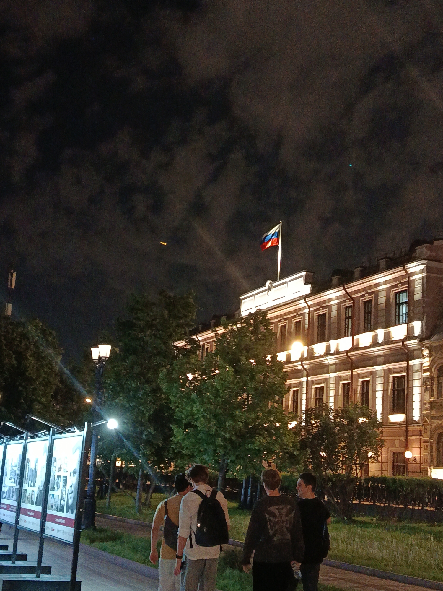 Captured at night, the image depicts a street scene in an urban environment, likely in Unknown, Unknown, dominated by a stately building illuminated with warm light, topped with the Russian flag. Four figures are walking on the sidewalk away from the camera. The background is filled with trees and street lamps. There are vertical poster displays on the left, and the sky is dark with visible clouds.