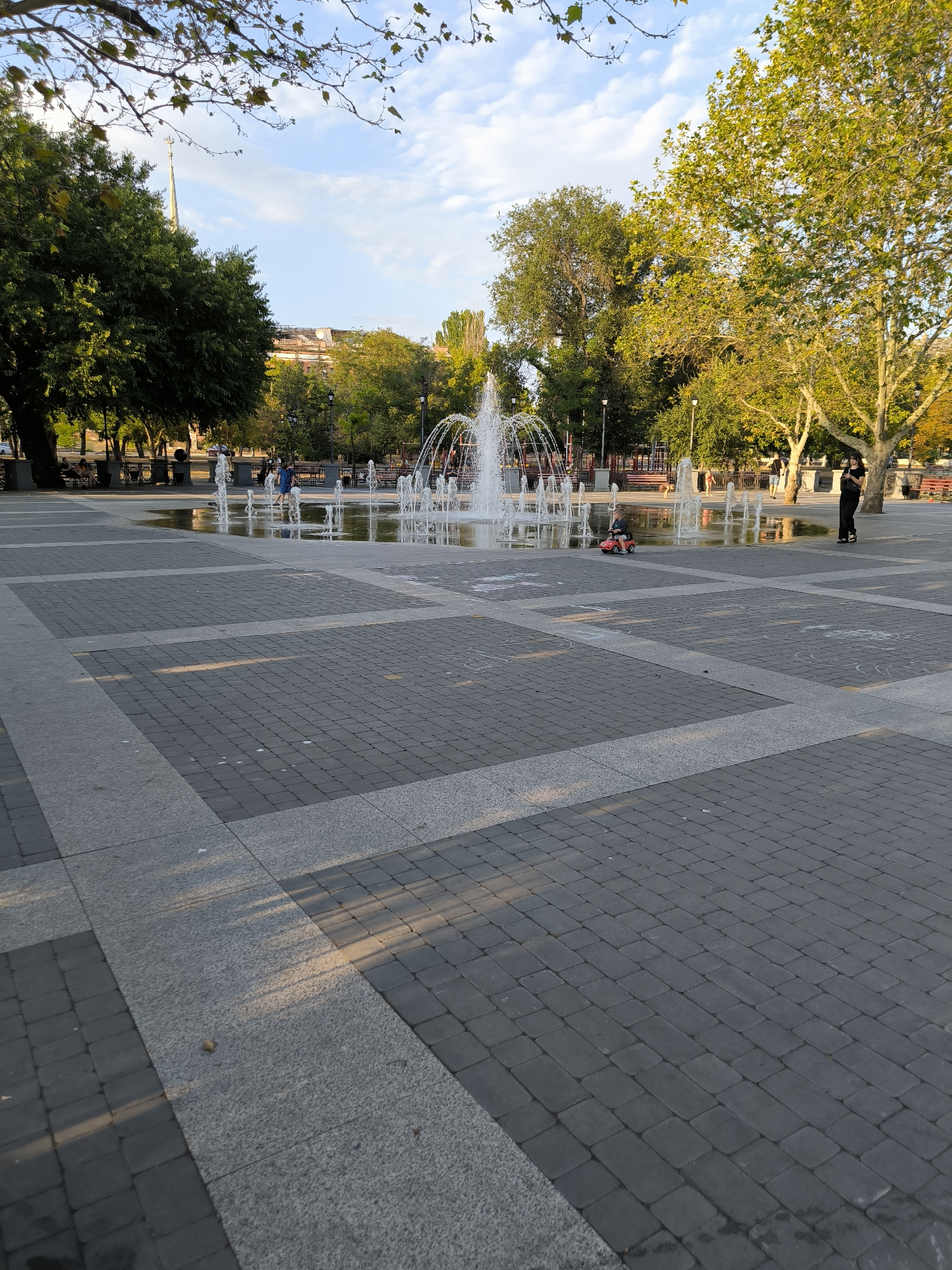 The image depicts a public square in Mykolaiv, Ukraine, featuring an interactive fountain in the center. The square is paved with dark gray rectangular stones, interspersed with lighter gray borders. Lush green trees with yellowing leaves surround the area, suggesting it is either late summer or early autumn. The sky is a mix of blue with scattered white clouds.
Several individuals are present in the scene. In the foreground, a child is seated in a small red toy car, which is on the paved ground near the fountain. Further back, a person stands near the fountain, looking at their phone. In the background, other individuals are seen sitting on benches or walking around the periphery of the square. The fountain itself consists of numerous water jets, creating arcs of water that reflect in the shallow pool formed on the ground. Streetlights and benches are visible throughout the square. The overall atmosphere appears relaxed and casual, suggesting a pleasant day in a city park.