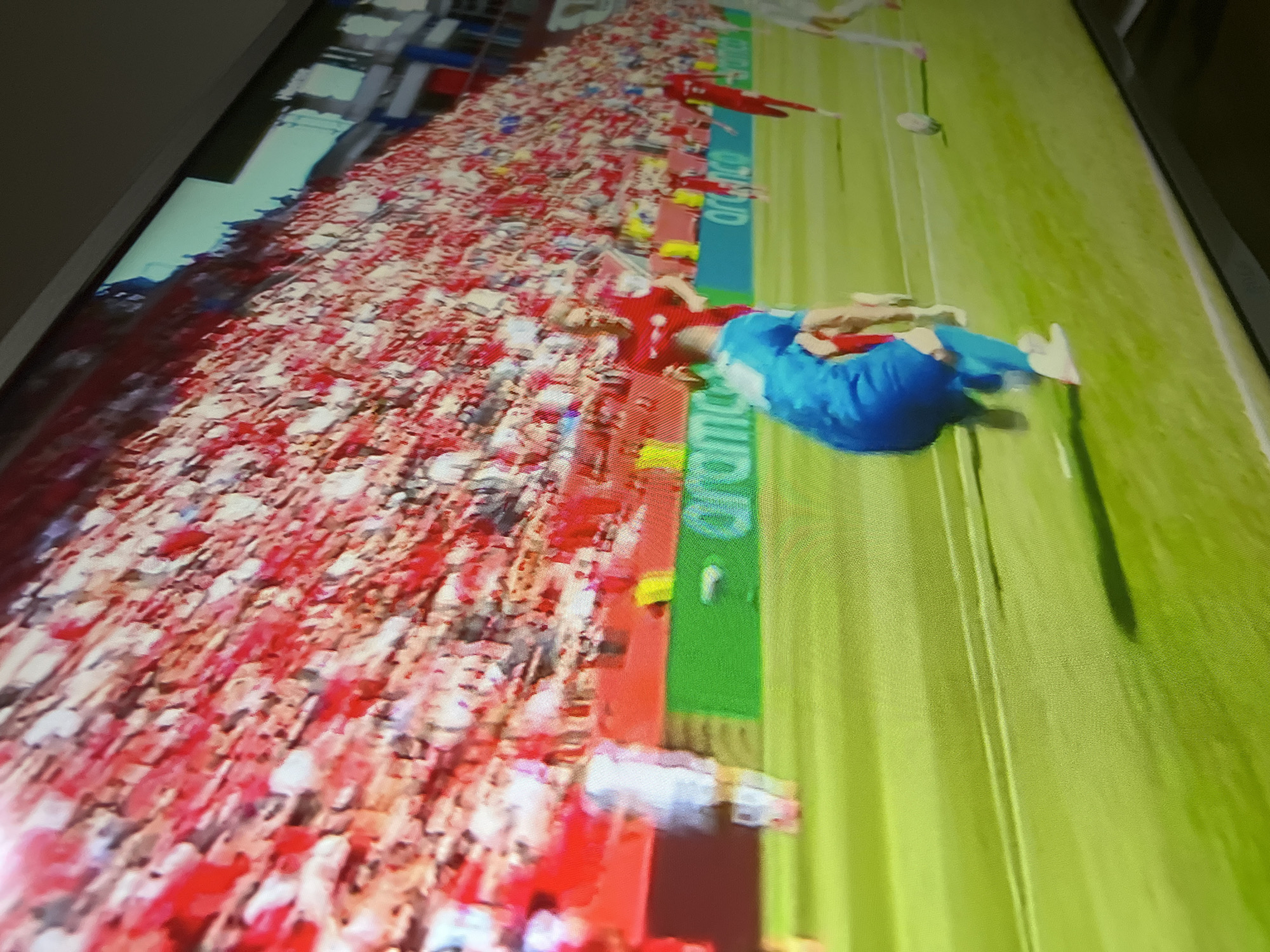This is a televised sporting event, likely a soccer match, viewed on a screen. A player in a blue uniform is running on a bright green field, with a soccer ball nearby. A crowd of spectators in red and white attire fills the stadium in the background. The scene appears to be captured at a slight upward angle, suggesting the camera is positioned below the action. The image quality is somewhat pixelated, typical of screen captures.