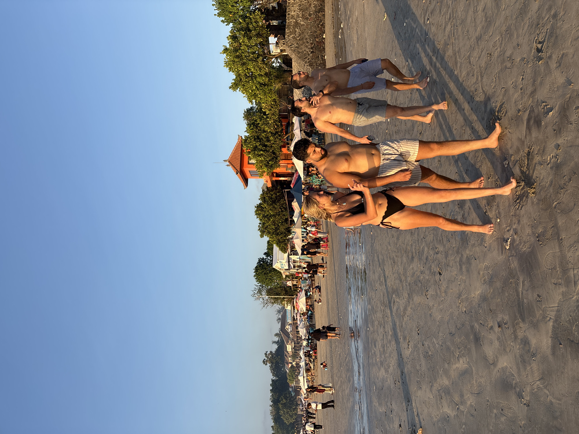 The image captures an outdoor scene on a sandy beach in Kuta Selatan, Indonesia, during the late afternoon or early evening, characterized by warm, directional sunlight casting long shadows. The sky above is clear and light blue.
In the foreground, four individuals walk abreast from left to right across wet, compacted sand marked with numerous footprints and scattered small rocks/shells. From left to right:
1. A blonde woman, wearing a black bikini, walks barefoot. She looks towards the person to her right, with her right arm slightly raised.
2. A man with dark hair and a beard, shirtless, wears light-colored striped shorts with a drawstring. He walks barefoot, looking downwards.
3. A man with dark hair and a beard, shirtless, wears light-colored striped shorts. He walks barefoot, looking to his left towards the previous man, with his left arm bent and hand near his chest.
4. A man with dark hair, shirtless, wears light blue shorts. He walks barefoot, looking to his left towards the previous man, holding a small dark object in his left hand.
The midground features a populated beach area with numerous individuals engaged in various activities. White, red, and blue beach umbrellas shade areas with tables and chairs. People are visible sitting, standing, and walking, including one person walking with a small child. The sand appears wet in patches, indicating proximity to the water's edge, though no substantial body of water is distinctly visible.
The background reveals a village or resort area situated behind the beach. Several single-story buildings with varied roofs (some white, some darker) are present, interspersed with abundant green foliage, including palm and leafy trees. A distinctive, two-tiered, pagoda-style orange building with traditional Indonesian architectural elements stands prominently on the right side of the background, partially obscured by trees. A stone wall is visible to the right of this building.