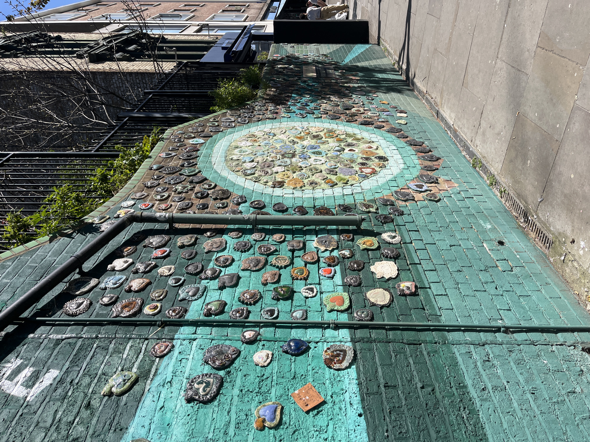 The image shows a green-painted brick walkway or alleyway in an unknown city. The bricks are painted teal, with vertical streaks of a darker green. A large mosaic circle is visible further down the walkway, composed of smaller, irregularly shaped tiles in shades of beige, blue, and green, surrounded by concentric circles of turquoise. Scattered across the brick surface are numerous small, circular or organically shaped objects, possibly ceramic or stone, with varying designs and colors. A black metal pipe runs across the bottom of the frame, close to the viewer, obscuring some of the bricks. On the left, a fire escape or stairway is visible, along with some vegetation, including small trees and climbing plants. A tall building can be seen on the left, and a taller, plain wall is on the right of the walkway. A person can be partially seen at the top of the walkway.