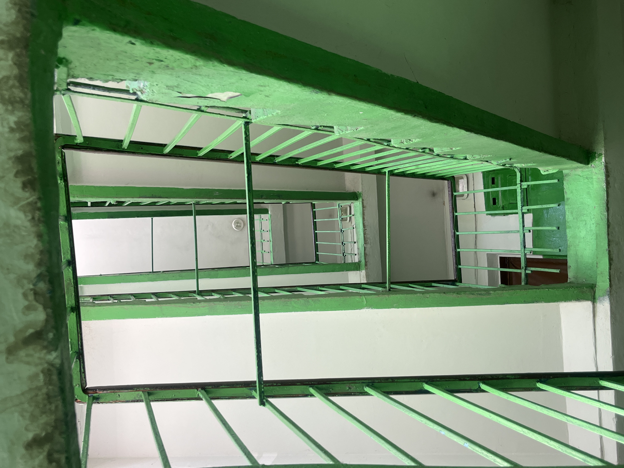 The image shows a view looking up a concrete stairwell with bright green metal railings. The stairwell appears to be in an apartment building, as suggested by the presence of doors visible on lower levels. The walls are painted a light color, and the concrete stairs are visible. The perspective is from a higher floor looking down into the well. There are no people or animals visible in the image. The overall impression is one of an old, utilitarian building, likely in an urban environment. The location of Drohobych, Ukraine is not visually discernible from the image itself.
