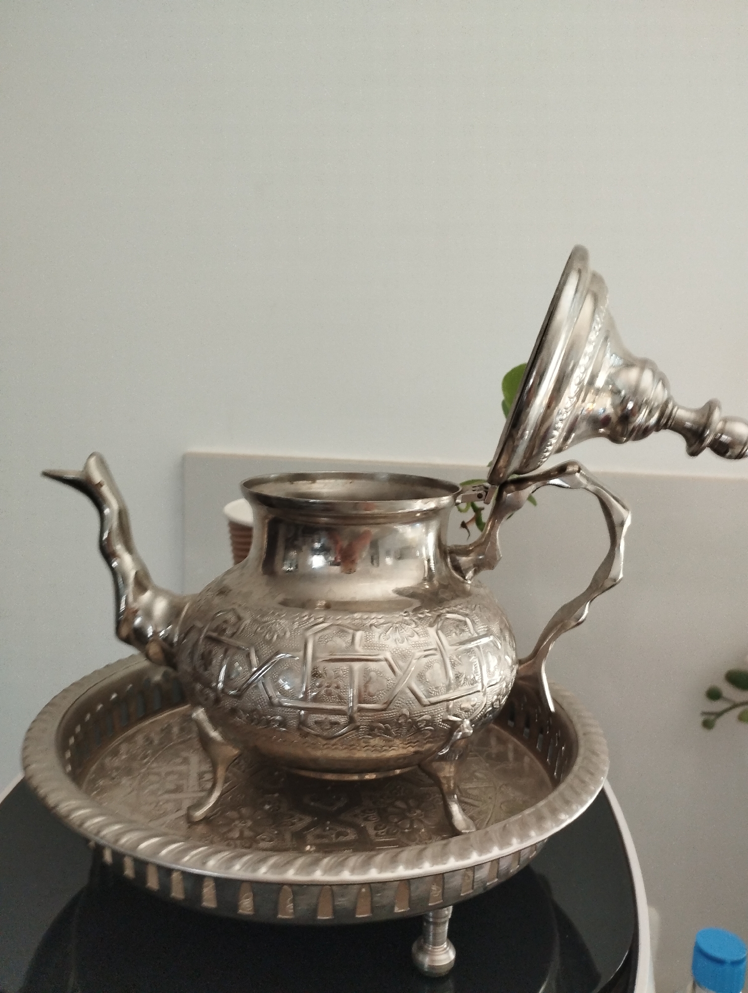 The image shows a vintage, ornate silver teapot on a matching silver tray. The teapot has a spout, a handle, and a lid that is slightly ajar, revealing a small green leaf inside. Both the teapot and the tray are intricately decorated with geometric patterns and embossed designs. The teapot is supported by three small feet. The tray also has decorative elements and a raised rim with a pattern of small openings. The setting appears to be indoors, with a plain light-colored wall in the background. There are some blurry green leaves from a plant visible on the right side of the image, suggesting a domestic environment. The lighting is soft and diffused, indicating it is likely daytime. No people are visible in the image. There is no discernible text in the image.