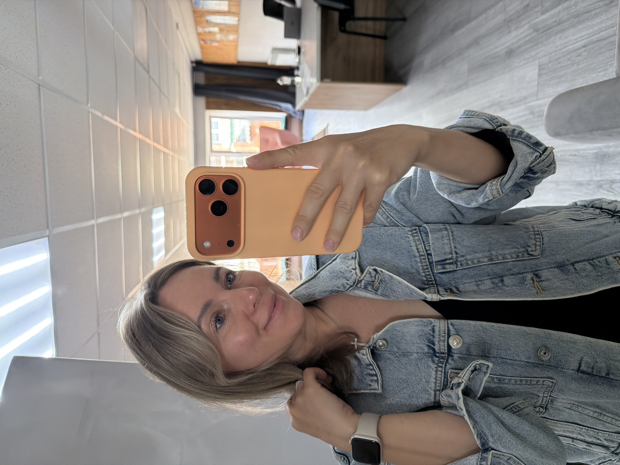 A woman with blond hair and blue eyes takes a selfie in what appears to be an interior space. She is wearing a light blue denim jacket over a black top and a silver cross necklace. A white smartwatch is visible on her left wrist, and she is holding an orange smartphone in her right hand, angled towards the camera. The background includes light-colored tiled walls, part of a window with sunlight streaming through, and glimpses of interior furnishings suggesting an office or living space. The photo does not explicitly indicate that the location is in the city of Chaikovsky, Russia.