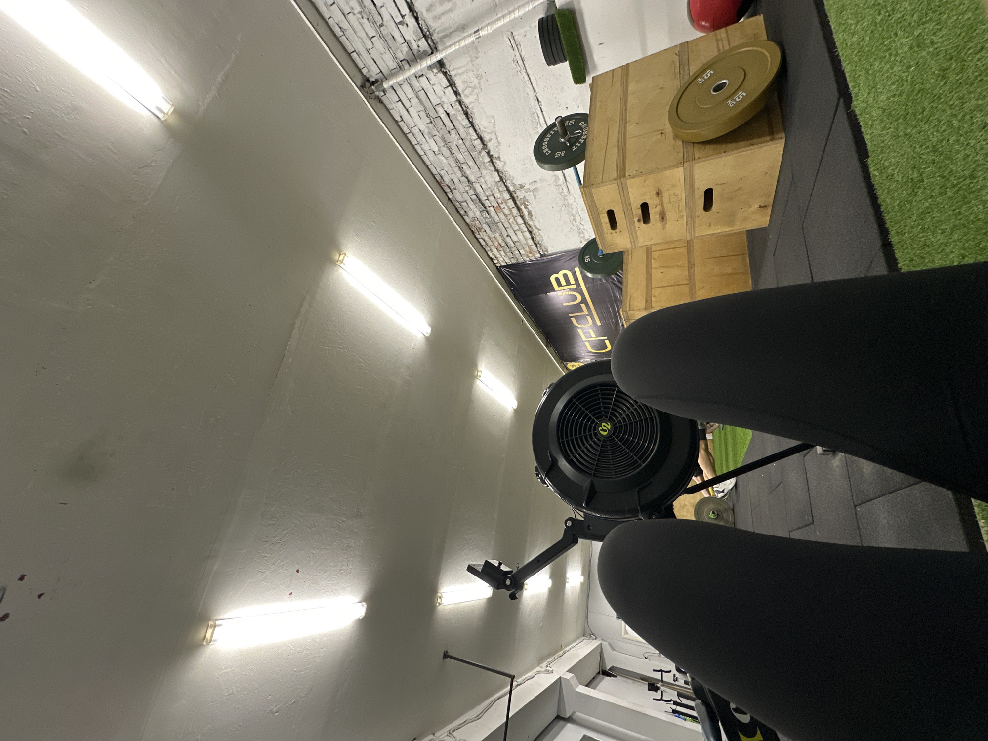 The image appears to be taken inside a gym, potentially in Chaikovsky, Russia given the presence of a sign with Cyrillic script that includes the letters "CFCLUB". The viewpoint is from a low angle, possibly first-person perspective of someone seated on a rowing machine. The focus is on the rowing machine and the person's legs, which are clad in black leggings. The surrounding gym equipment includes stacked wooden boxes with weight plates on top, a barbell with weights, and artificial turf. The gym has a concrete ceiling with fluorescent strip lighting. The walls are painted white with exposed brickwork in some areas. The overall lighting is bright.