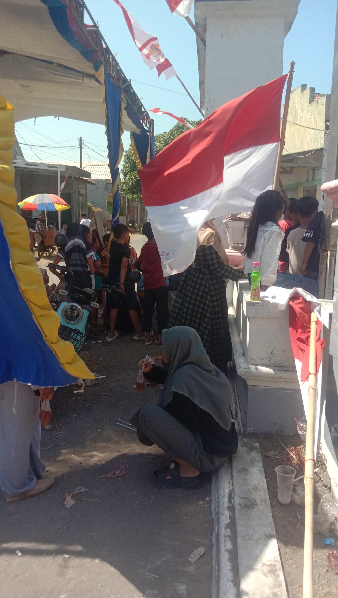This is a medium-shot, outdoor, vertical photograph taken on a sunny day in Karanganyar, Indonesia. The scene appears to be an event or gathering, with several people present. In the foreground, a person wearing a grey hijab and dark clothing is crouching, holding a small clear bag with red liquid. To their right, a plastic cup is on the ground next to a bamboo pole. In the middle ground, a light blue moped is parked. A group of people, some appearing to be children and teenagers, are standing around the moped and in the background. Indonesian flags, red and white, are prominently displayed and waving in the wind. There are also blue and yellow fabric decorations overhead, suggesting a temporary setup. The environment is somewhat urban, with buildings and power lines visible in the background. The ground is a mixture of dirt and concrete.