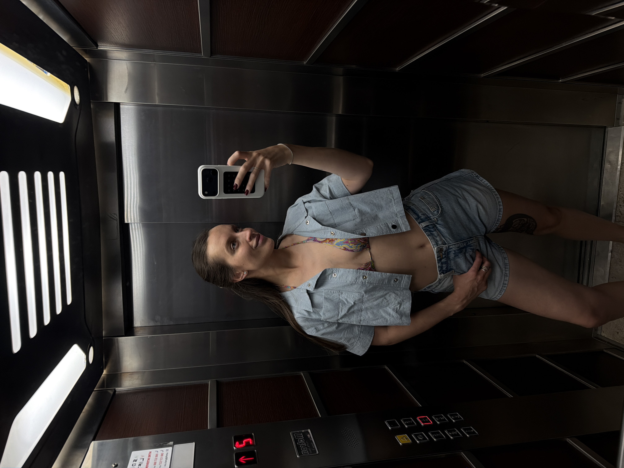 The image is a photograph taken in an elevator. A woman is centered in the frame, holding a smartphone with a white case in her right hand, presumably taking a selfie. She has long brown hair and wears a light blue, short-sleeved button-down shirt over a patterned bikini top and denim shorts. A tattoo is visible on her left thigh.
The elevator's interior is modern, featuring stainless steel walls and a dark wood panel beneath the control panel. The control panel displays the number "5" illuminated in red. To the left, the elevator door shows a set of vertical lights on the edge.
The photograph provides insufficient information to identify the precise location in Budva, Montenegro.