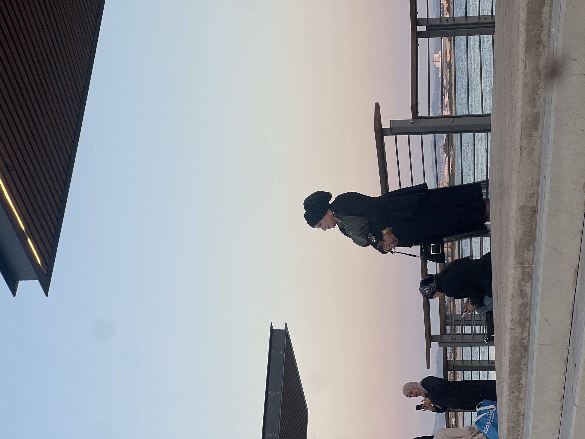 The image captures an outdoor scene at what appears to be a promenade or pier in Acre, Israel, during either sunrise or sunset, indicated by the soft, gradient sky transitioning from light blue to pale pink and orange hues. The perspective is slightly angled, suggesting the photo was taken from below or to the side.
In the mid-ground, a woman wearing a dark jacket, a green top, and a black head covering (possibly a turban or beanie) is leaning forward, looking down towards something out of frame. She has golden bracelets on her right wrist. Behind her, another individual in dark attire and a patterned head covering also bends forward, engaged with something below the railing. Further right, closer to the viewer, a woman wearing a light-colored hijab and a dark abaya is looking at her mobile phone. A blue and white shopping bag, partially visible at her feet, bears Hebrew text, including the word "שוק" (market) and parts of what might read "משפחת דוד" (David's family).
The backdrop features a metal railing, beyond which a calm body of water (the sea) stretches out, with distant land and buildings visible on the horizon. To the far left, two dark, angular architectural structures rise against the sky; one is part of a building with vertical slats, and the other is a distinct, dark, triangular element. The foreground on the right side is dominated by a thick concrete barrier or wall, running parallel to the railing. The overall atmosphere is serene and contemplative, with individuals enjoying the view or engaged with personal devices.