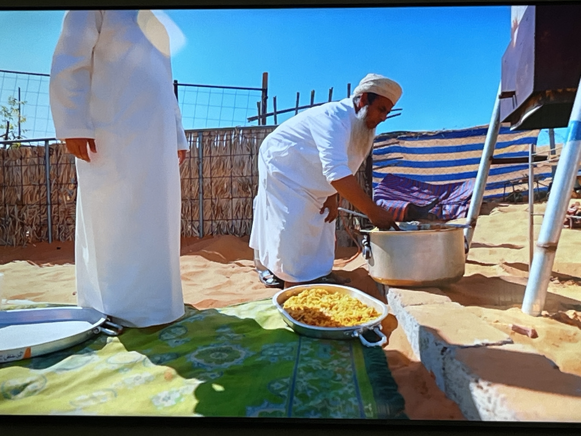 Two men in traditional white clothing stand in a sandy outdoor environment. One man, facing away from the camera, stands near a large metal pot on a raised structure. The second man, facing the pot, is in the process of adding something to it. In the foreground, a metal dish filled with yellow food sits on a green and white patterned cloth. The background shows a fence, some vegetation, and a blue and striped fabric structure. The environment appears to be a desert or arid region. There is no indication in the image that this scene is located in Kharkiv, Ukraine.