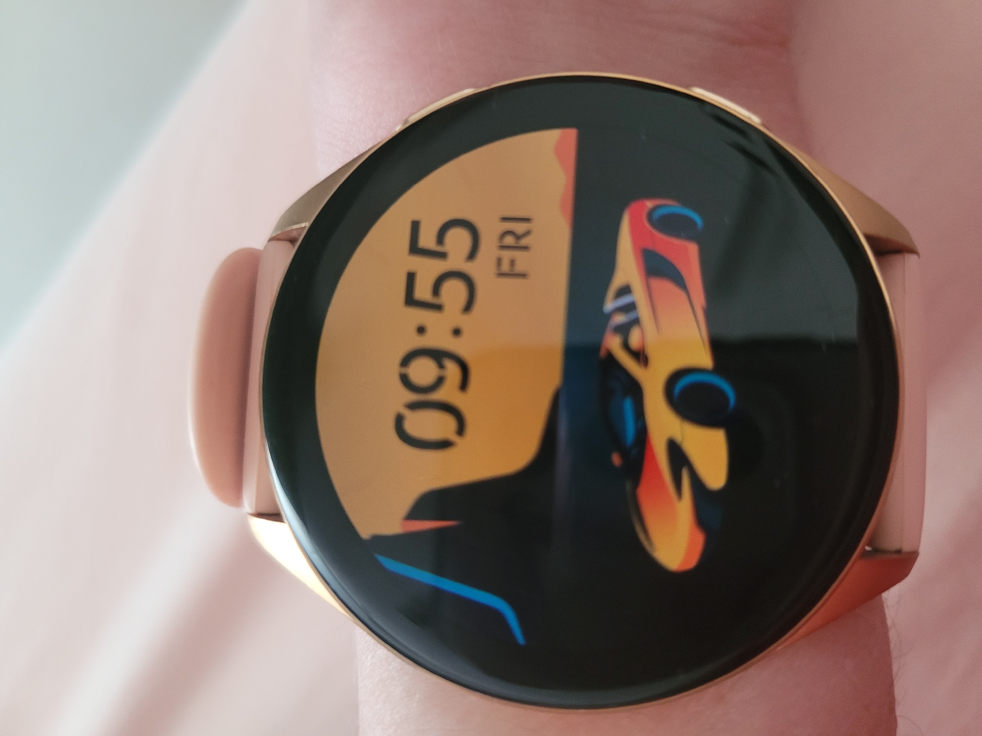 The image shows a close-up view of a smartwatch with a digital watch face. The watch face displays the time as 09:55 and the day as FRI. The background of the watch face features an illustration of a stylized car in orange and yellow, with blue accents, set against a sunset-colored landscape with abstract mountain shapes. The smartwatch has a rose gold colored bezel and is worn on a person's wrist, which is partially visible. The background is a soft pink fabric, likely clothing or a blanket. There are no discernible elements in the image that would allow for the determination of its location in Kyiv, Ukraine.