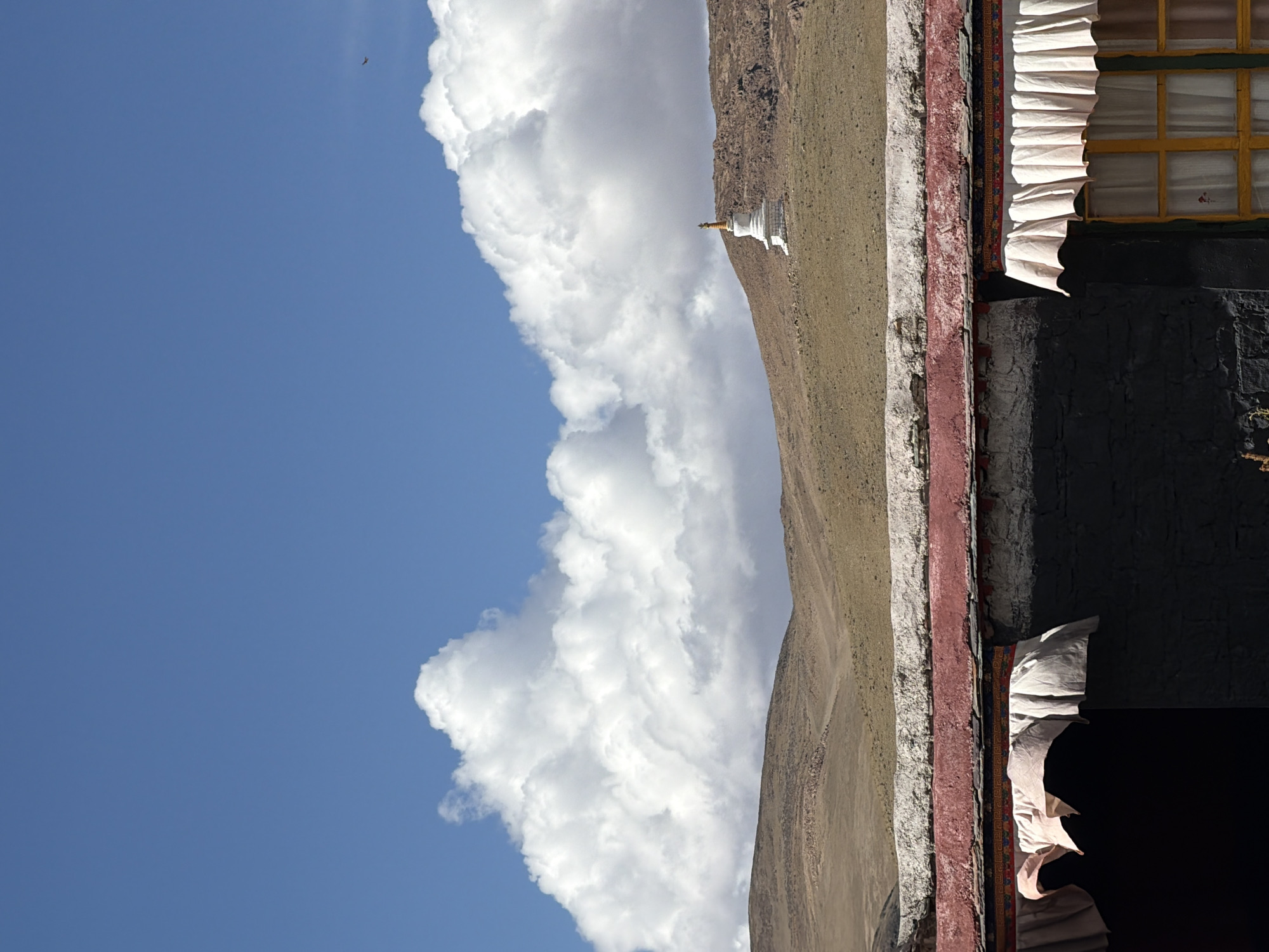 This striking image, captured in Labrang township, China, presents an expansive landscape under a bright, daytime sky. The scene is composed of a vibrant blue sky, a vast barren mountain range, and a section of a traditional building, depicted as if viewed with a horizontal horizon.
The upper portion of the image is filled with a clear, deep blue sky, punctuated by large, dramatic cumulus clouds. These voluminous white clouds, brilliantly lit by the sun, cast gentle shadows upon themselves, creating a sense of depth. A tiny, dark bird is visible soaring in the upper left of the sky.
Below the sky, the arid, golden-brown slopes of a mountain stretch across the mid-ground. The terrain appears rugged and sparsely vegetated, characteristic of high-altitude environments. Perched on one of these slopes, a distinctive white stupa (chorten) with a shimmering golden spire stands as a focal point, indicating a site of spiritual significance.
The lower right quadrant of the image shows a close-up of a building, exhibiting architectural features common to the region. Its sturdy walls are painted in sections of dark grey, rich red, and lighter off-white/grey. A window with a warm yellow frame is visible, partially draped by a pleated, white textile hanging. Another patterned textile, possibly a decorative banner, is layered vertically between different colored wall sections.
The photograph captures a moment of serene grandeur, highlighting the blend of natural beauty and cultural heritage under bright, clear weather conditions. No people or visible text are present.