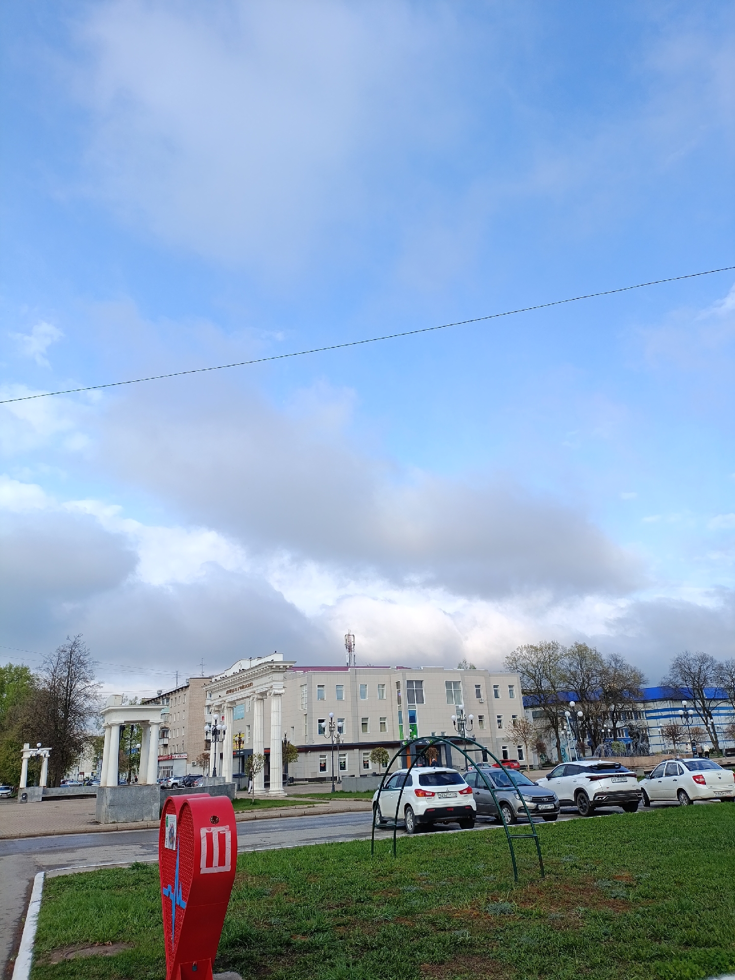 The image shows a view of Chaikovsky, Russia, on a partly cloudy day. In the foreground is a green grassy area with a red, perforated metal structure. Behind the grass and a paved road, a row of parked cars is visible. In the background, a building with white columns stands, along with a white arched structure to the left. Overhead, a blue sky is interspersed with gray and white clouds. A thin cable stretches across the upper part of the frame.