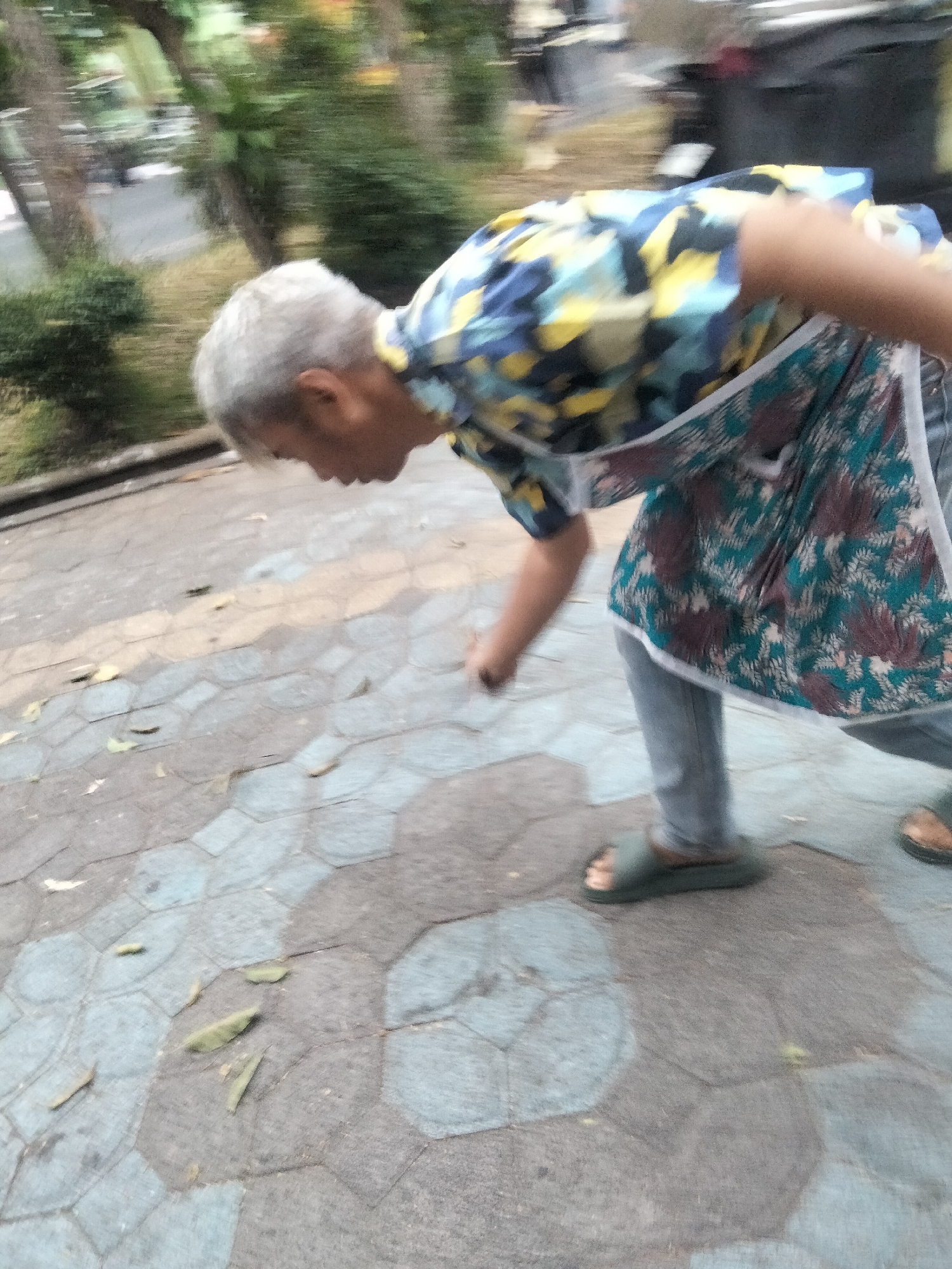 In Surakarta, Indonesia, a person with gray hair and wearing a patterned shirt and apron is bending down to pick up something on a hexagonal patterned pathway. The person is wearing blue jeans and dark sandals. The pathway appears to be made of interlocking stone or concrete tiles. There are some fallen leaves on the pathway. In the background, there are trees and greenery, suggesting an outdoor setting, possibly a park or public walkway. The image is blurred, indicating movement.
