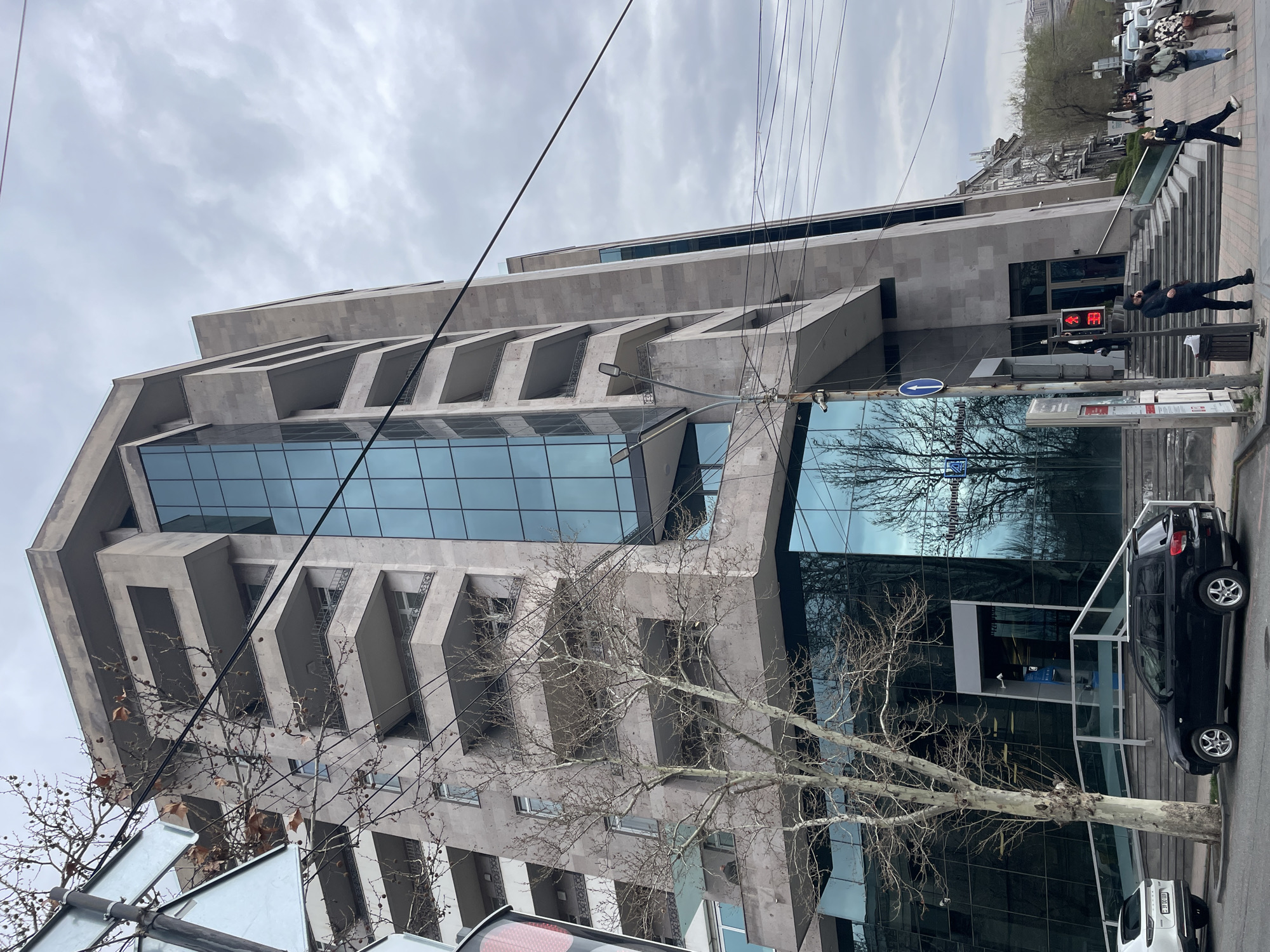 The image depicts a modern building in Yerevan, Armenia, constructed from light-colored stone and glass. The building features protruding rectangular balconies and large glass windows, reflecting the surrounding environment, including a leafless tree.
In the foreground, a tree without leaves stands alongside a city street. A black car is parked near the building’s entrance, next to stairs leading up to it. A traffic light is visible, showing a red light. Pedestrians are on the sidewalk beyond the building, some walking up the stairs. Overhead, power lines crisscross the sky. The sky is overcast with grey clouds. Street signs are visible on poles near the traffic light.