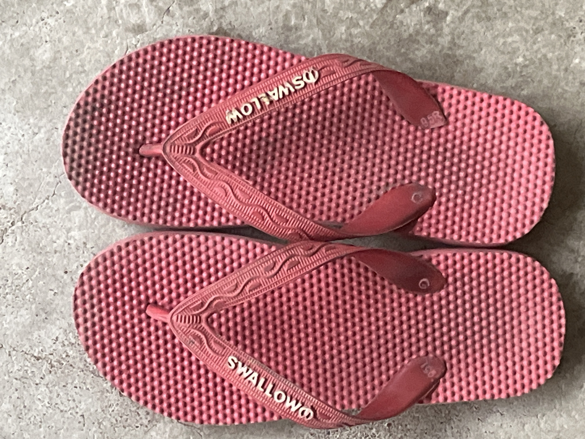A pair of pink flip-flops with the word "SWALLOW" embossed on the straps lies on a textured, light-colored concrete surface. The flip-flops are positioned side-by-side, with the left flip-flop slightly overlapping the right. The surface texture of the flip-flops features a grid of small raised bumps. The concrete surface appears worn and has a grayish hue with some darker markings. The lighting suggests an outdoor environment.