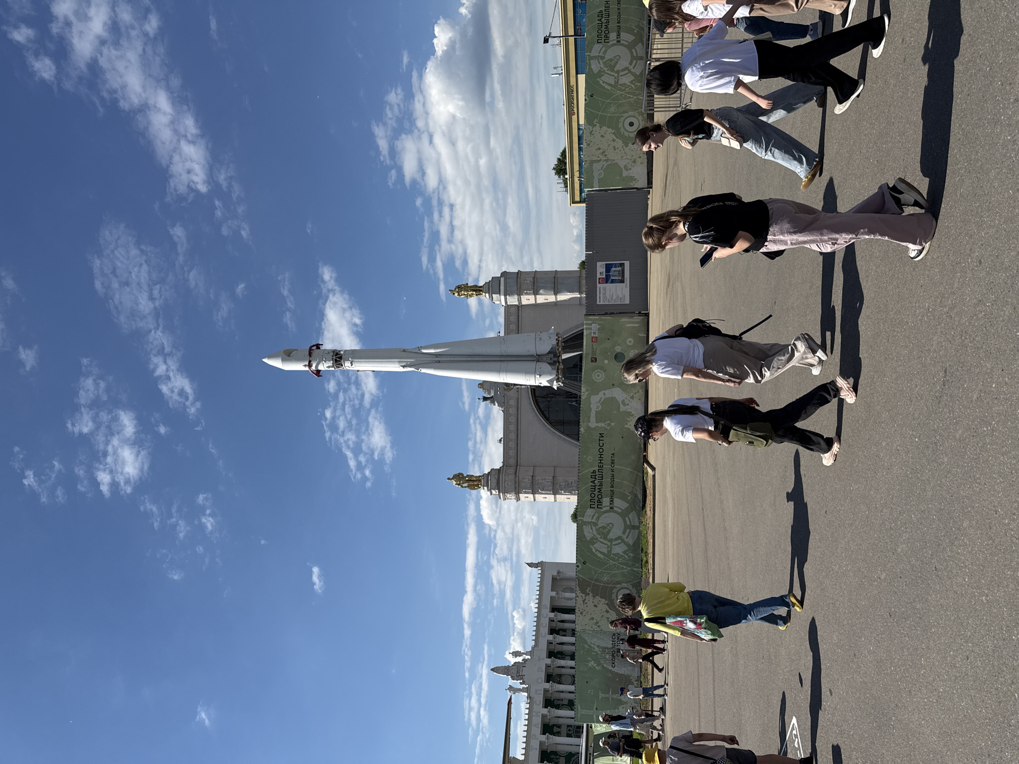 This image, taken on a bright, sunny day, depicts a bustling outdoor scene featuring a towering rocket and ornate buildings, with several people walking along a paved pathway. The perspective is tilted, showing the ground and subjects vertically.
The primary subject is a large white rocket, likely a replica or monument, standing tall against a clear blue sky dotted with scattered white clouds. Its multi-stage structure and pointed nose cone are clearly visible. Behind the rocket and to its right, several grand, light-colored buildings with intricate details and golden decorative elements on their spires or domes add architectural splendor to the setting.
On the right side, a group of young adults is casually walking along an asphalt path. Their long, distinct shadows stretch across the ground, indicating a high sun position, likely mid-day or early afternoon. Some individuals are looking at their phones, while others appear to be observing their surroundings. Green banners or hoardings line the pathway, displaying text and graphic designs.
Visible text on the banners includes "ПЛОЩАДЬ ПРОМЫШЛЕННОСТИ" (Industrial Square), "В ТАНЦЕ ВОДЫ И СВЕТА...." (In the dance of water and light...), and "СКОРО ЗДЕСЬ" (Coming Soon Here), suggesting an ongoing or future development or event. The word "РОСКОСМОС" (Roscosmos) is also discernible, reinforcing the space theme, along with "ВДНХ" (VDNKh). This abbreviation strongly indicates the location is the Exhibition of Achievements of National Economy in Moscow, which contradicts the provided context of Khimki, Russia.
