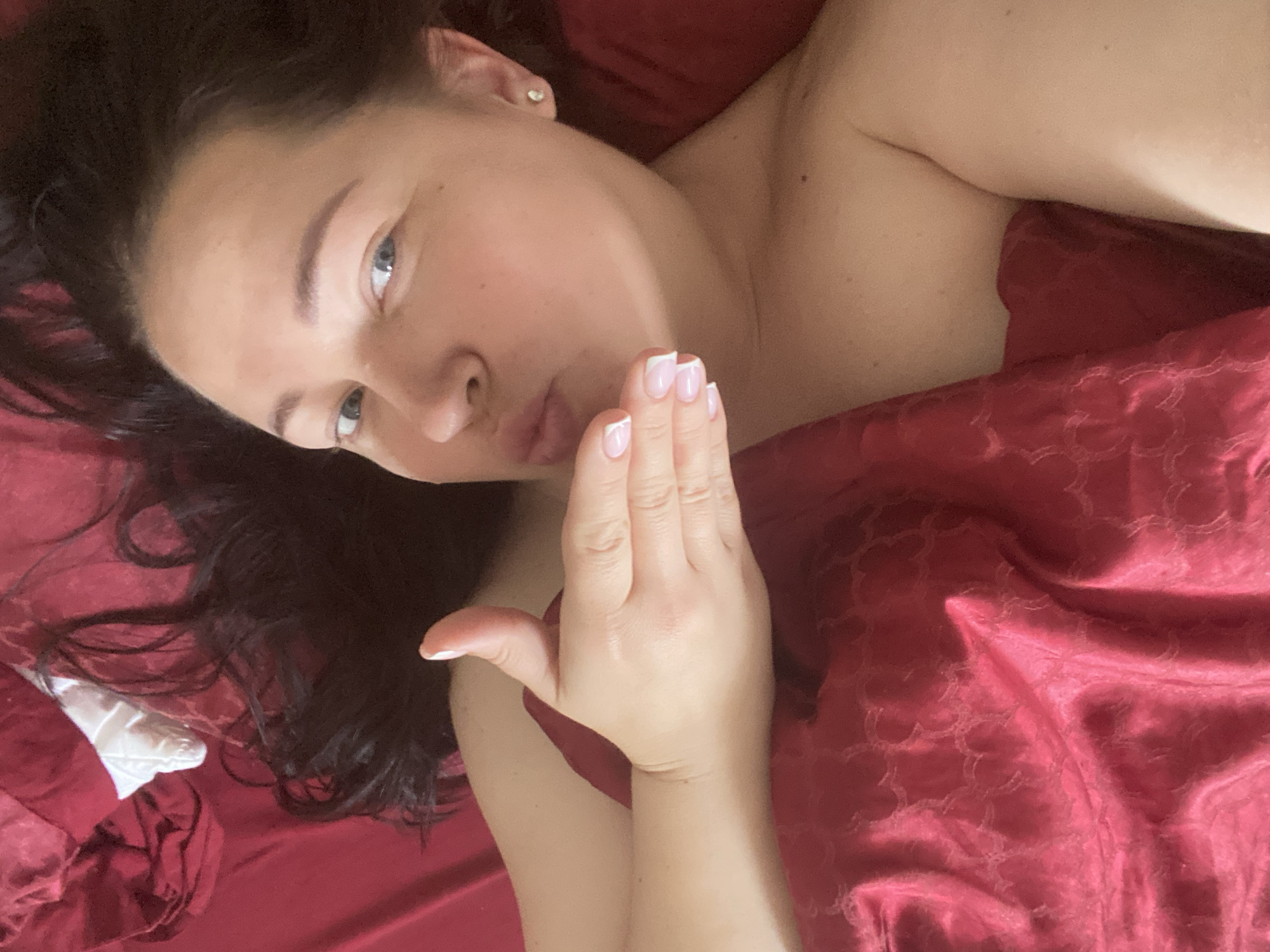 A woman lies in bed, partially covered by a red satin sheet. She looks at the camera, her dark hair spread out on the red bedding. She has light skin, blue eyes, and is making a kissing gesture with her lips. Her right hand is raised, with her fingers facing the camera, showing a French manicure. The woman is wearing a small earring in her left ear. The location of Sochi, Russia, cannot be determined from the image.