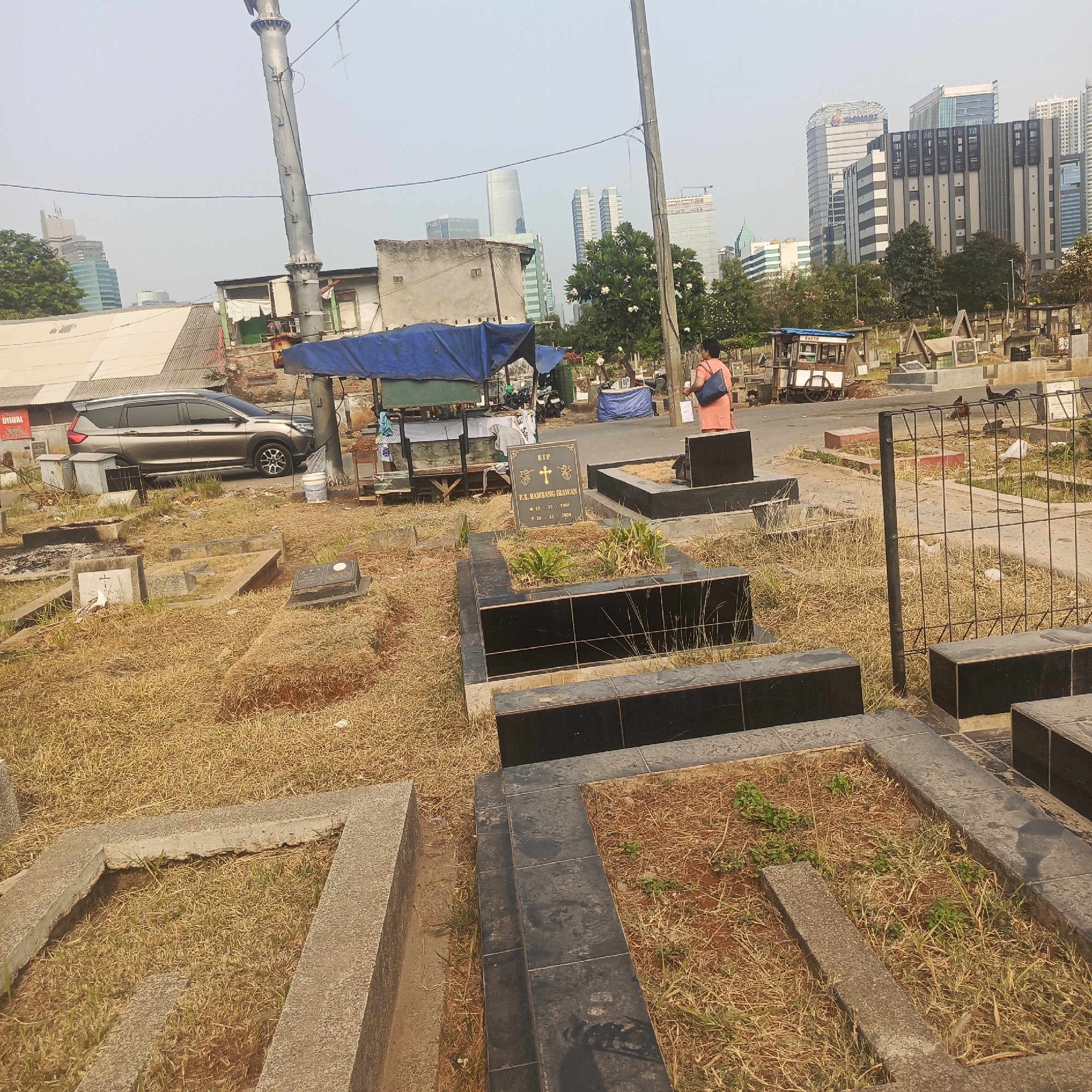 The image captures a daytime scene in a cemetery in South Jakarta, Indonesia. The foreground is dominated by graves, some with concrete borders and others with black tile coverings. Dry, yellow grass covers most of the ground, interspersed with a few small green plants. A gravestone with the inscription "RIP F.K. HAMBAGO IRAWAN" and dates "05 - 11 - 1966" and "20 - 11 - 2020" is visible in the mid-ground.
In the background, a woman in an orange top and blue bag walks away from the camera. A street vendor cart with the word "BAKSO" is parked on the asphalt road. Beyond the cemetery, a modern city skyline rises, featuring tall buildings, including ones with "XL SMART" signage. A silver SUV is parked on the left side of the image, near some low-rise structures with blue tarps. Overhead, utility poles and wires crisscross the sky, which appears hazy. The overall impression is a juxtaposition of a traditional cemetery against a backdrop of urban development.