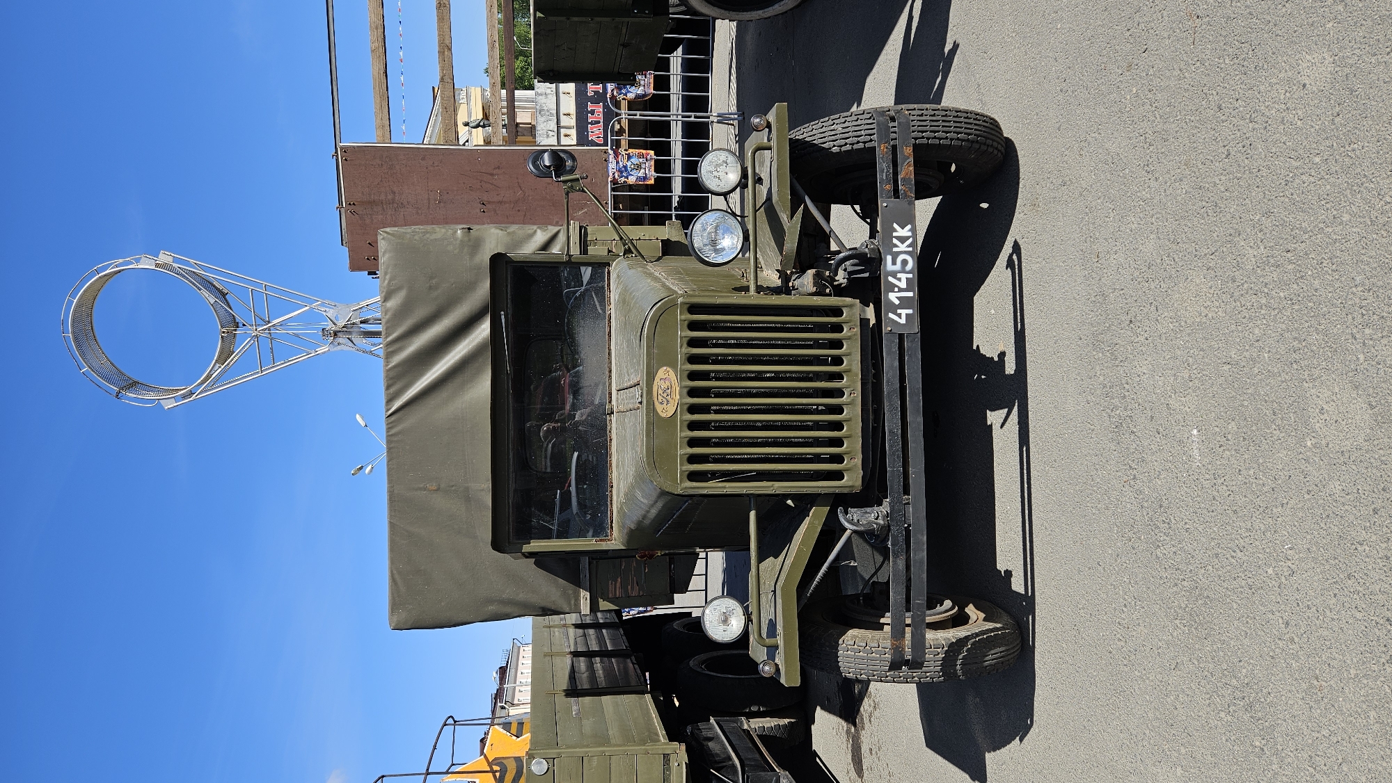 A vintage olive green truck is parked on an asphalt surface under a clear blue sky. The truck has a canvas cover over its rear bed. A metal grille is visible on the front of the truck, with a circular emblem above it. Two round headlights are mounted on the front fenders. A black license plate with white lettering "41-45KK" is attached to the front bumper. To the left of the truck, part of another green truck is visible. In the background, there is a tall, white, circular metal structure and a brown building. Several people are reflected in the truck's windshield.