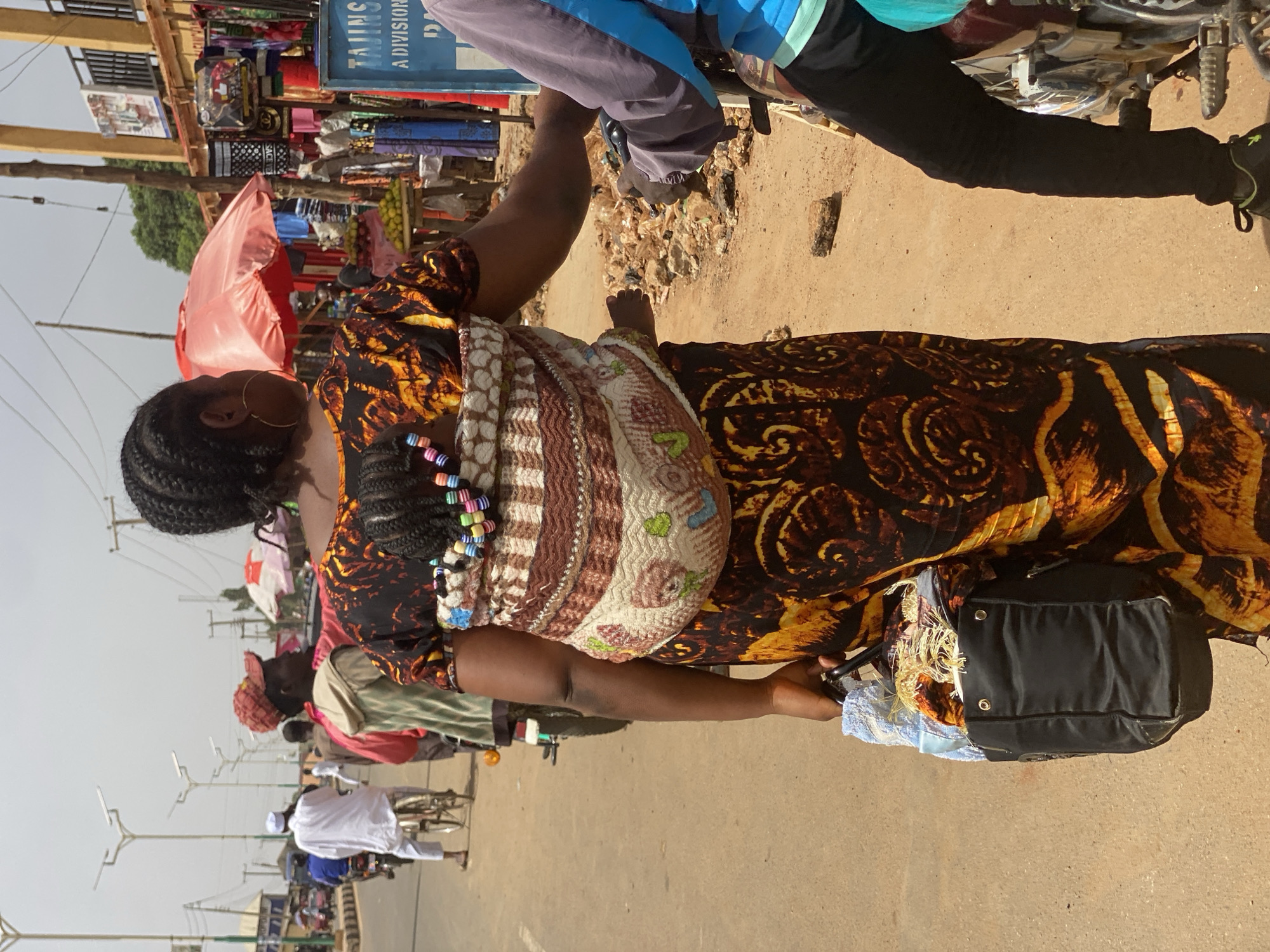 The image depicts a bustling street scene in Kafanchan B, Nigeria, on a bright, sunny day. The primary subject is a woman walking in the foreground, viewed from her left side. She has dark braided hair adorned with colorful beads and is wearing gold hoop earrings. She is dressed in a vibrant, patterned gown featuring swirls of orange, brown, and black. A baby is securely carried on her back in a patterned fabric wrap, with one small foot visible. The woman carries a black handbag in her left hand.
Behind her, to the right, a person is seated on a motorcycle, wearing black pants and a blue and light blue top. The ground is a sandy-brown dirt road with some debris. To the left of the woman, the street is lined with market stalls or shops, indicated by makeshift structures and various goods on display, including what appears to be clothing and other items. A red umbrella provides shade in this area.
Further down the street, several other people are visible, some riding bicycles, and one person in white attire. Modern-looking street light poles extend along the road. In the background, a blue sign is partially visible, showing the words "TINGS" and "A DIVISION". The overall scene captures daily life and commerce in a lively urban or semi-urban setting.