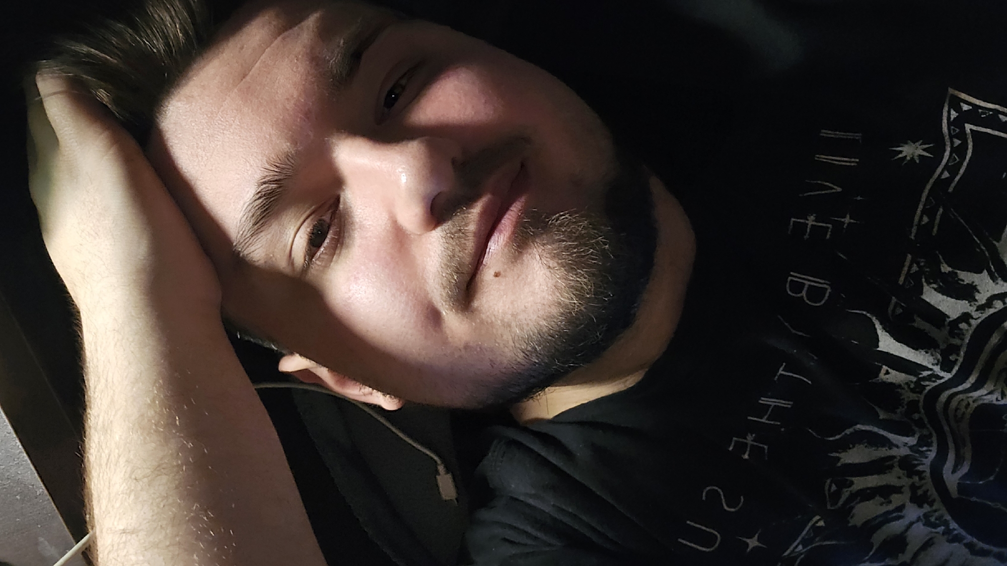 A close-up photograph shows a man lying down. His head rests on his left hand, which is raised slightly. The man has short brown hair and a beard. He's wearing a black t-shirt with white writing and patterns. The lighting is dim, focusing on his face. There is no indication in the image of the location being Sofia, Bulgaria or any other specific place.