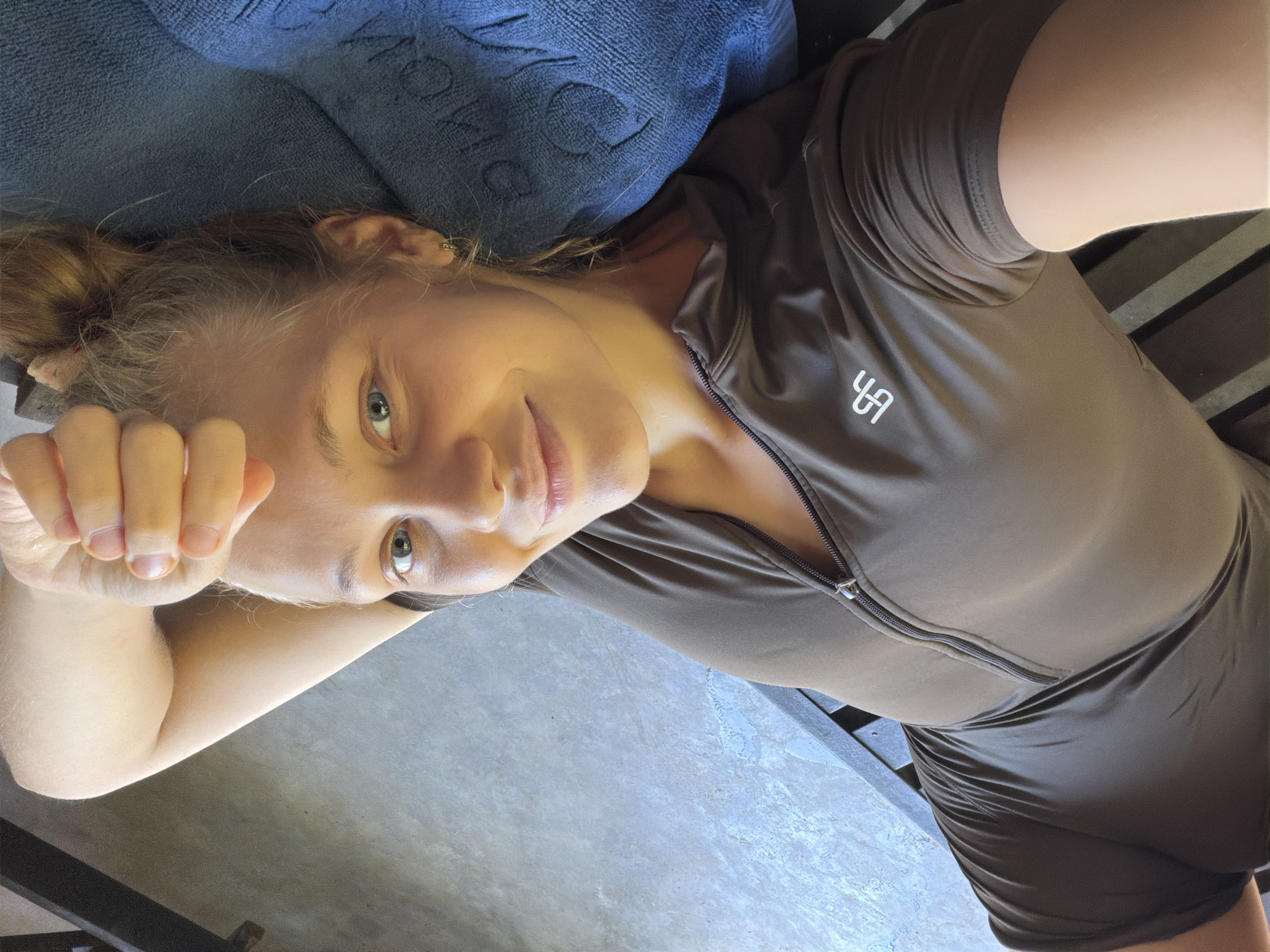 The media file is a close-up, self-portrait of a young woman lying down. She has blonde hair pulled up in a bun and is looking at the camera with blue eyes. Her right arm is bent at the elbow and resting behind her head, with her hand near her face. She is wearing a short-sleeved, brown zip-up top with a white logo on the chest. The background consists of a blue textured fabric, possibly a towel or blanket, and a concrete floor with a slatted wooden surface. The lighting suggests it could be daytime. The woman's expression appears relaxed and content. There is no visible text other than the logo on her shirt. The location is specified as Tay Nha Trang Ward, Vietnam.