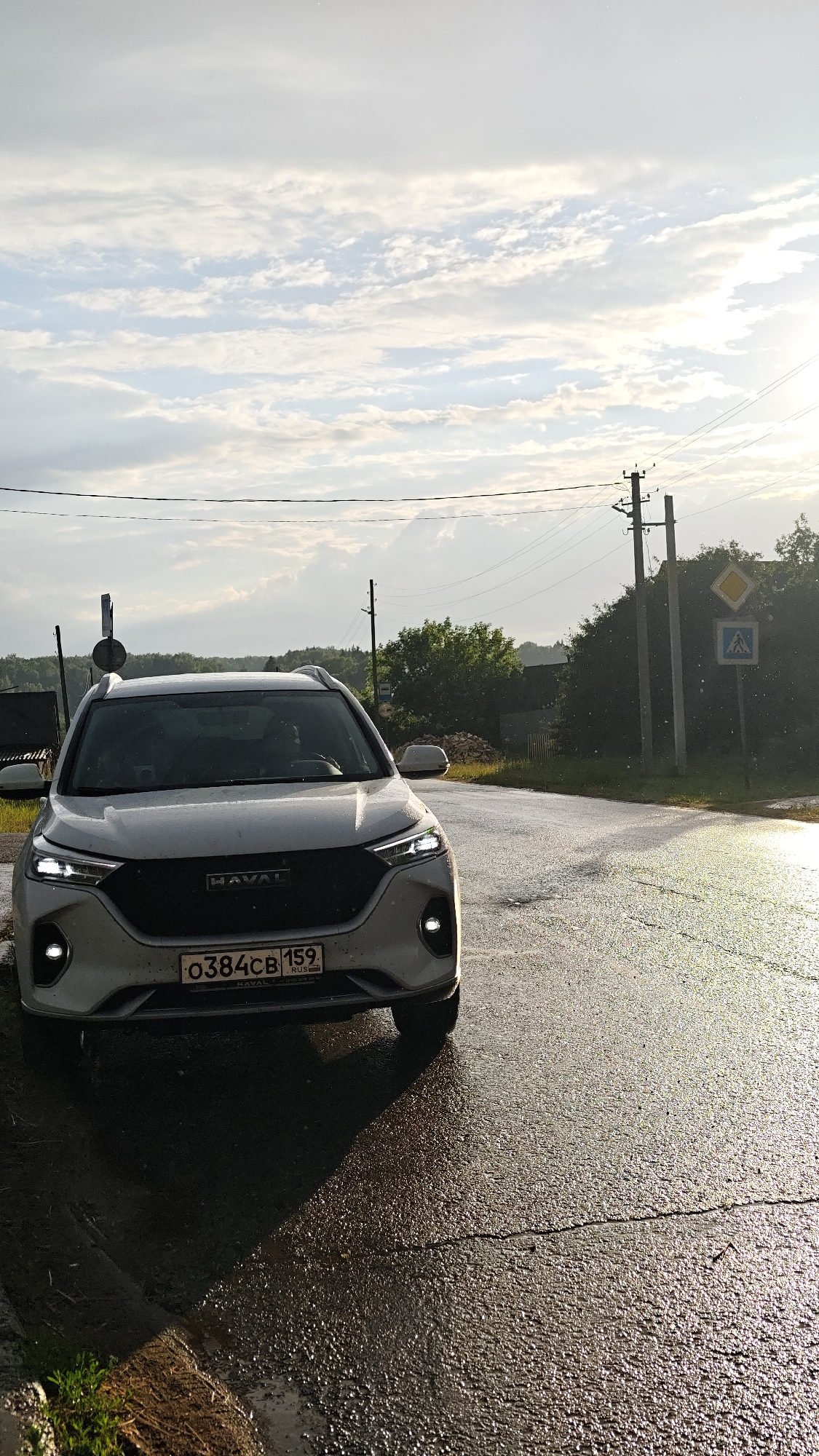 A white Haval SUV is parked on the side of a wet, asphalt road. The car's headlights are on, illuminating the scene. The road surface reflects the overcast sky, with streaks of sunlight breaking through. The car has a license plate that reads "0384CB 159 RUS".
The setting appears to be a rural area in Уральское, Russia. In the background, there are trees, power lines, and utility poles. A yellow diamond-shaped sign with a blue pedestrian crossing symbol is visible on a utility pole to the right of the car, suggesting a road intersection or a designated crossing area. The weather seems to be damp, possibly after rain, given the wet road and reflections. The sky is filled with a mix of clouds and patches of bright sky, indicating it might be late afternoon or early evening, with the sun attempting to break through. No people are visible in the image. The overall mood is serene and perhaps a bit melancholic due to the weather and the stillness of the scene.