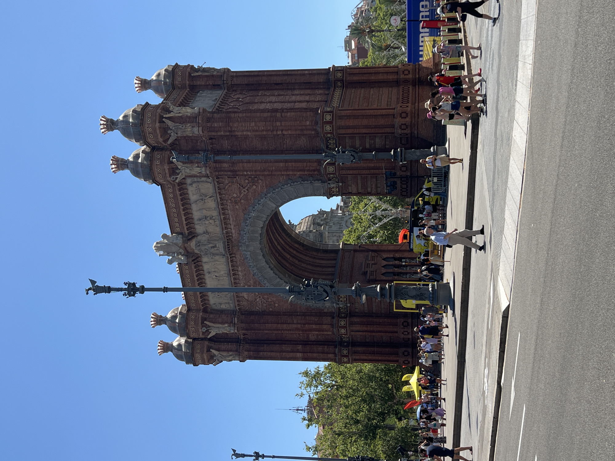 The image shows a wide shot of the Arc de Triomf in Barcelona, Spain, on a clear, sunny day. The monumental arch, constructed of red brick, is the central focus, with a large arched opening in the center. Decorative elements adorn the top of the arch, including what appear to be sculptures and urn-like structures topped with red finials. A lamppost with a decorative base and a bird-like sculpture on top stands in the foreground to the left of the arch.
In front of the arch, a street or promenade is visible with a crosswalk. Several people are walking and gathering in this area. Some individuals are crossing the street, while others are standing in groups, possibly tourists or event attendees. Various tents and banners are set up behind the crowd, suggesting a possible event or festival. A yellow van is parked to the left, and further back, trees and buildings can be seen. The sky is a bright, cloudless blue. The overall scene is one of a public space in a city, bustling with activity on a warm day.