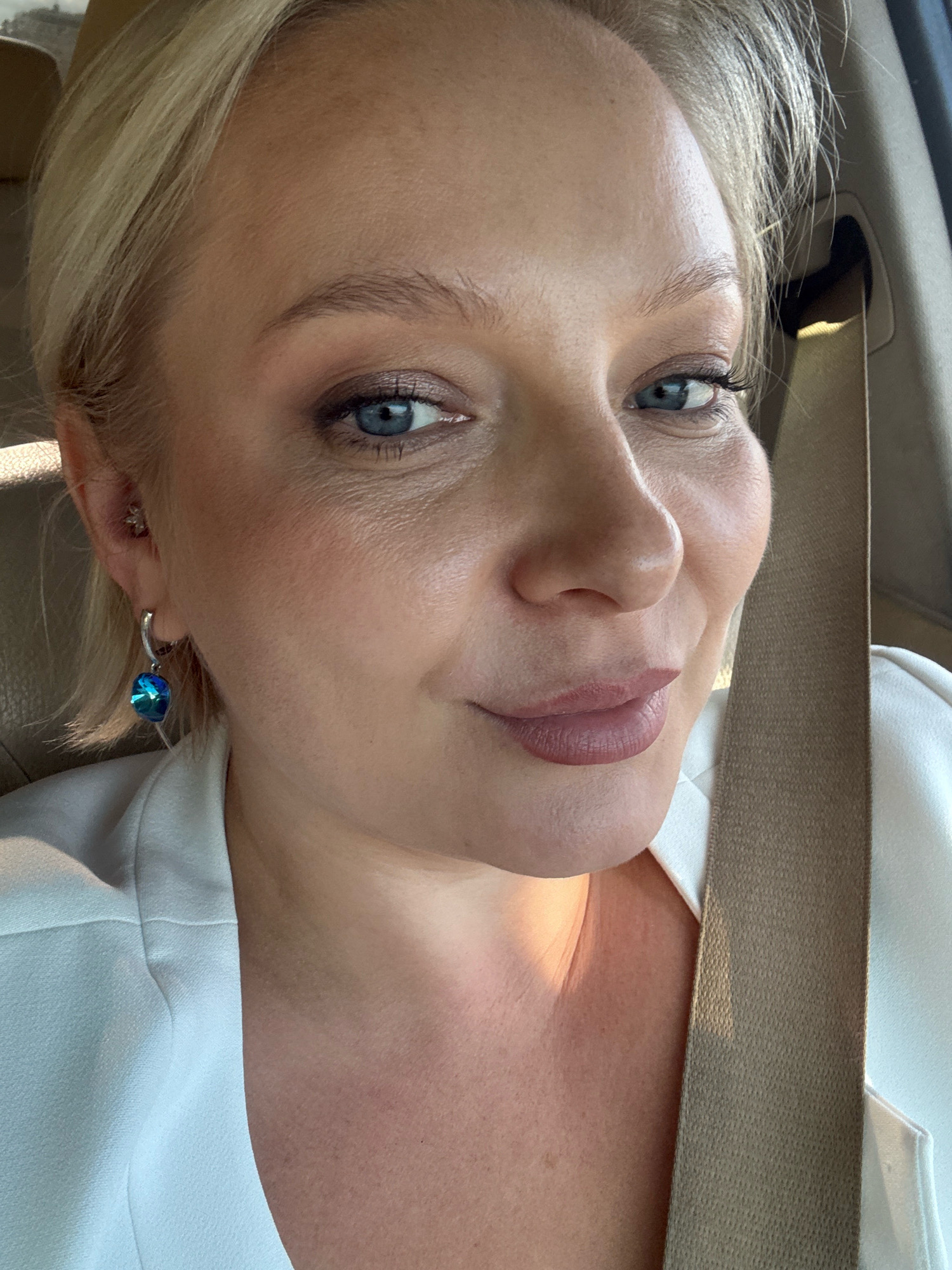 A blonde woman with blue eyes and fair skin is shown in a close-up, selfie-style shot. She is wearing a white blazer and a large, blue, teardrop-shaped earring. Her hair is short and styled to the side. The background appears to be the interior of a vehicle, with a seatbelt visible. The lighting suggests an indoor setting, possibly daytime. No specific location within Лядскі сельскі Савет, Belarus can be determined from the image alone.
