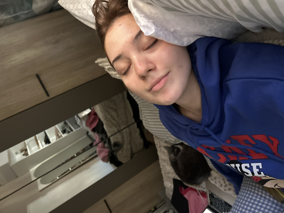 The image displays an indoor scene within a residence located in Schiltigheim, France. A young female is depicted resting on her right side in a bed, her eyes closed. She has light skin and brown hair, and is wearing a blue hooded sweatshirt with partially visible red and white text. Her head rests on a white pillow with subtle grey stripes. To her immediate left, a dark grey cat is curled up and resting on the bed. A section of a dark-colored laptop keyboard and a pink garment are visible on the bed surface near the cat. The background features a wardrobe with mirrored panels. The reflection in these mirrors reveals another part of the room, showing a bed with light-colored bedding, upon which various items of clothing are loosely placed, and open shelving containing multiple pairs of shoes.