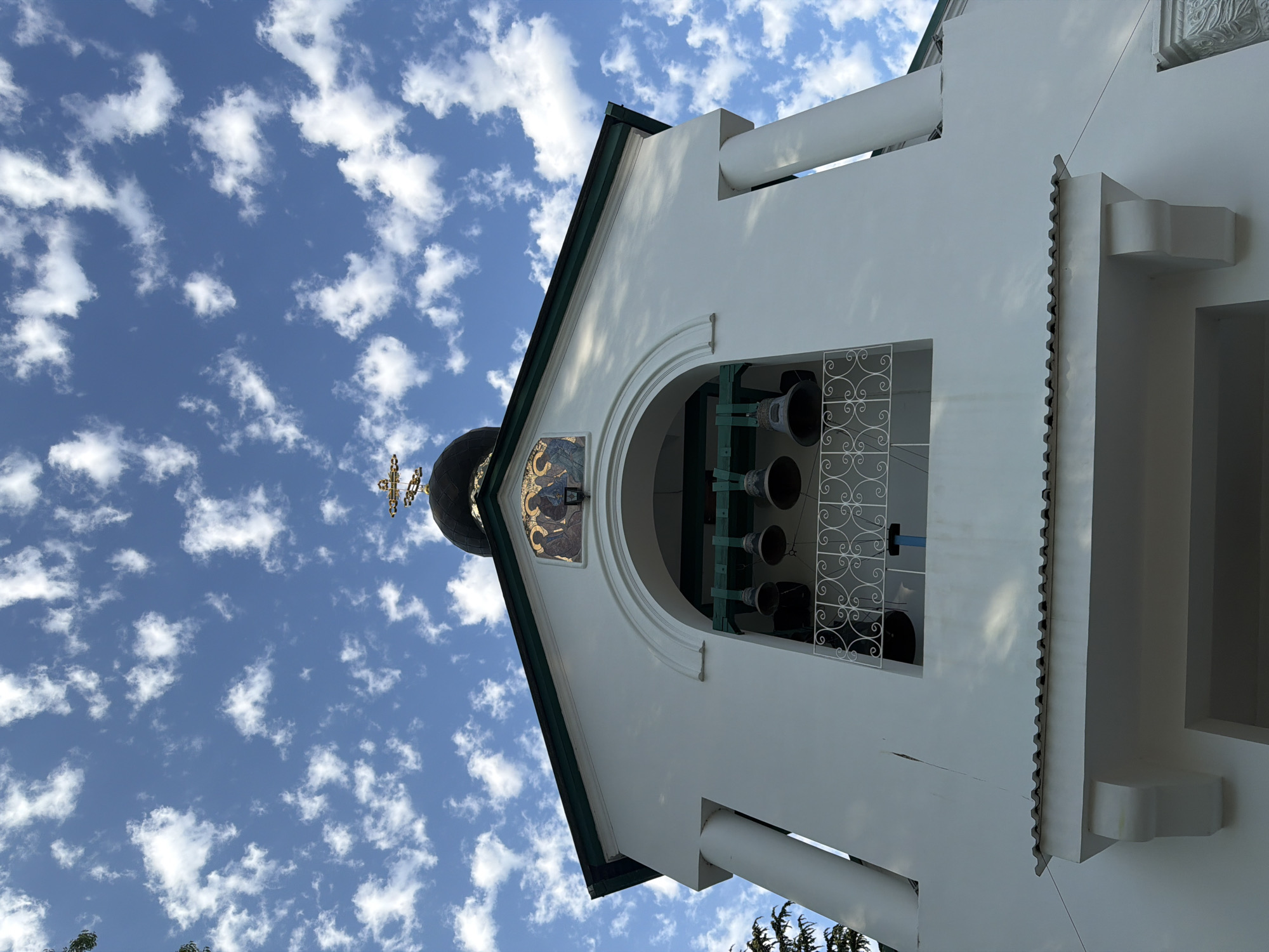The image shows a close-up, low-angle view of a white bell tower, likely part of an Orthodox church, against a bright blue sky filled with scattered white clouds. The tower has a green-tiled roof and features an arched opening where several bells are hung. Ornate metalwork is visible in the window, and a small mosaic depicting religious figures is mounted above the arch. A dark dome topped with a cross is partially visible to the left of the bell tower. The sunlight suggests it is daytime, and the clear sky with clouds indicates fair weather. The style of the architecture, particularly the dome and religious iconography, points to a Russian Orthodox church. There is no visible text in the image. The scene is serene and evokes a sense of spiritual architecture against a natural backdrop.