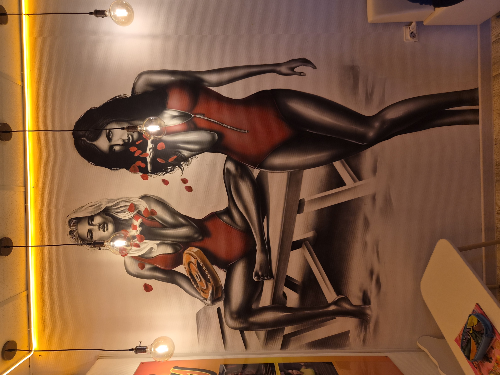The image shows a wall mural in what appears to be an interior setting in Sochi, Russia. The mural depicts two women in red swimsuits, rendered in a stylized, almost airbrushed manner. One woman has dark hair, the other blonde. Both are depicted with scattered red rose petals around them. The women are sitting on lifeguard stands, also rendered in the same style. Light fixtures, consisting of bare Edison-style bulbs on long cords, are positioned in front of the mural, visually intersecting with the women's faces. A bright yellow neon light is present along the left edge of the frame. The background color of the wall is off-white. In the lower right of the frame, parts of tables and a poster can be seen.