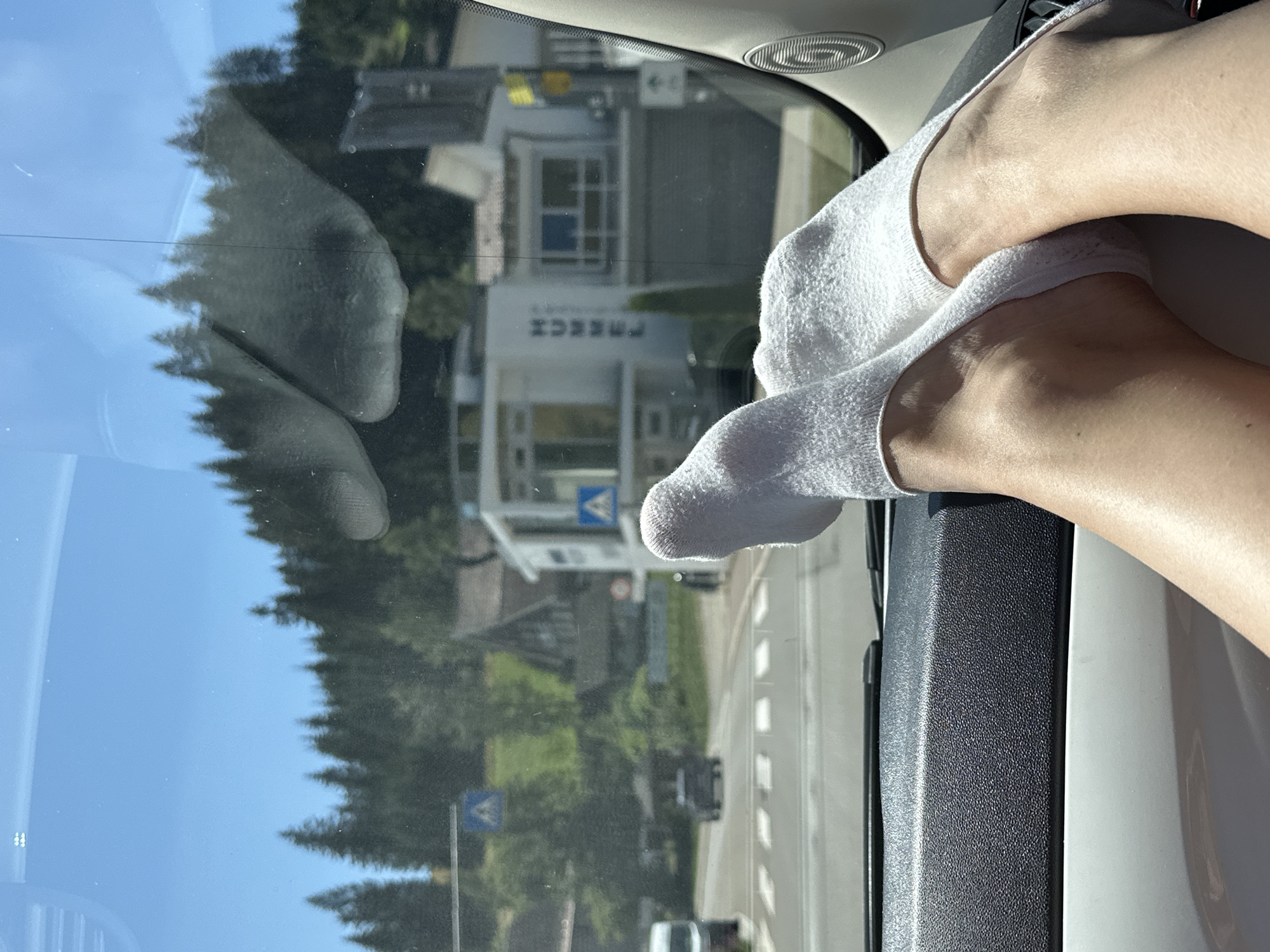 The image depicts a person's feet, clad in white ankle socks, resting on the dashboard of a car. The car's interior, specifically the textured dashboard and a portion of the passenger door panel, is in the foreground. Through the windshield, a street scene is visible in Furtwangen im Schwarzwald, Germany. The view includes a pedestrian crossing, traffic lights, and several buildings, one of which has the word "HUKSE" visible on its facade. Trees and a hillside form the background, under a clear blue sky. The overall impression is of a person traveling by car on a sunny day in a German town.